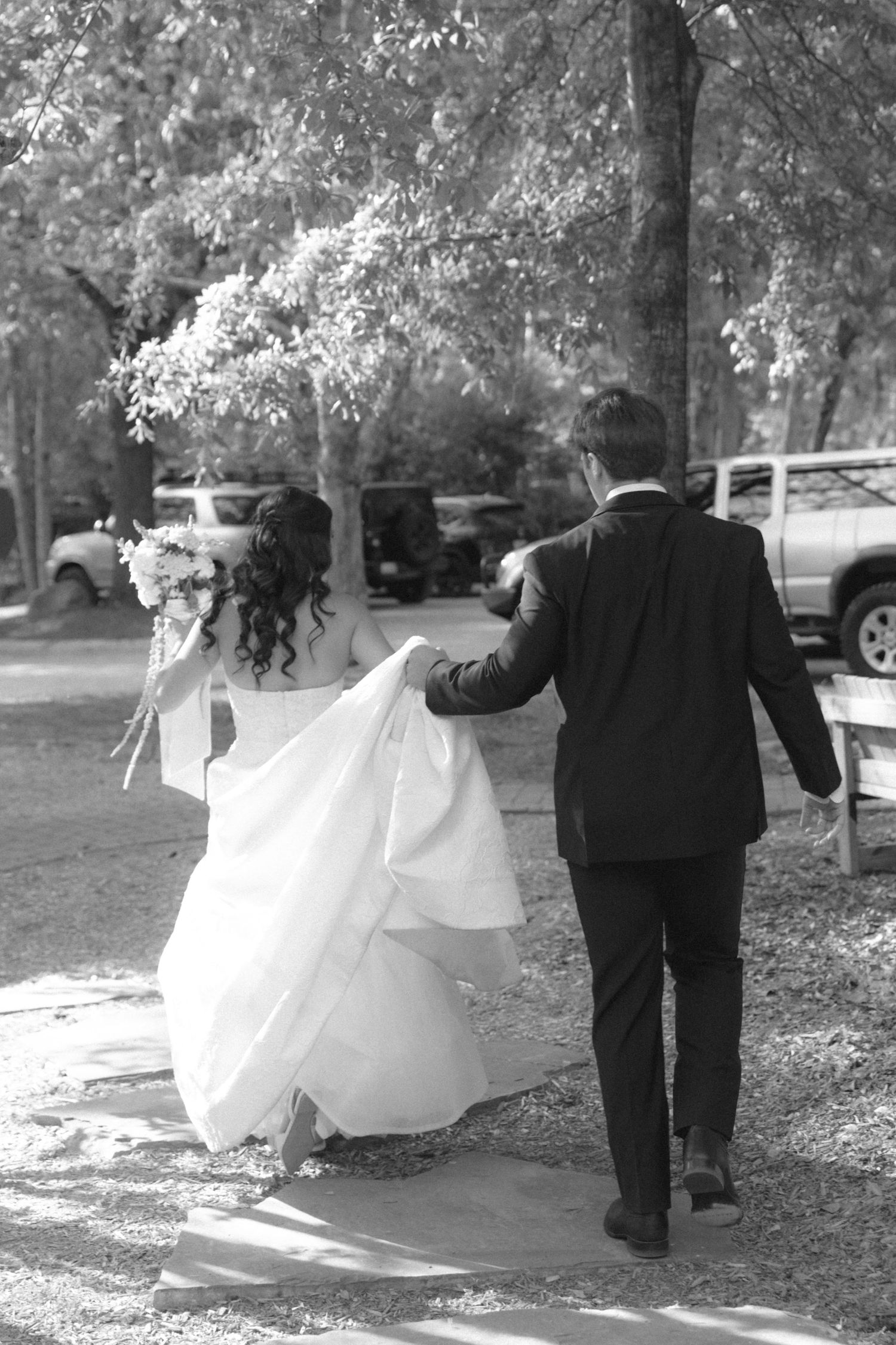 Black and white photo of bride and groom walking away, groom holding bride's white wedding gown train.