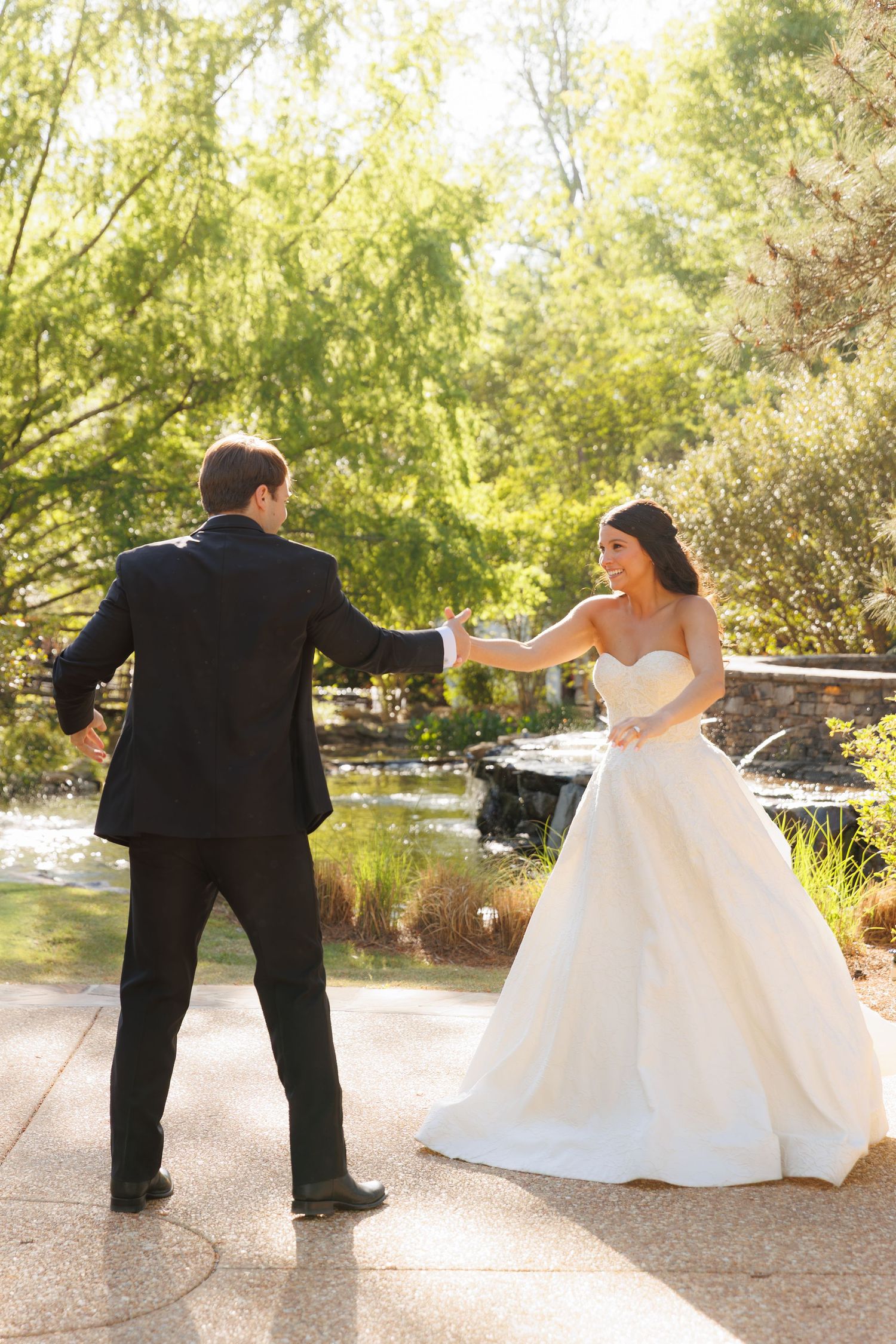 Bride in white gown and groom hold hands dancing outdoors in a sunlit garden setting.