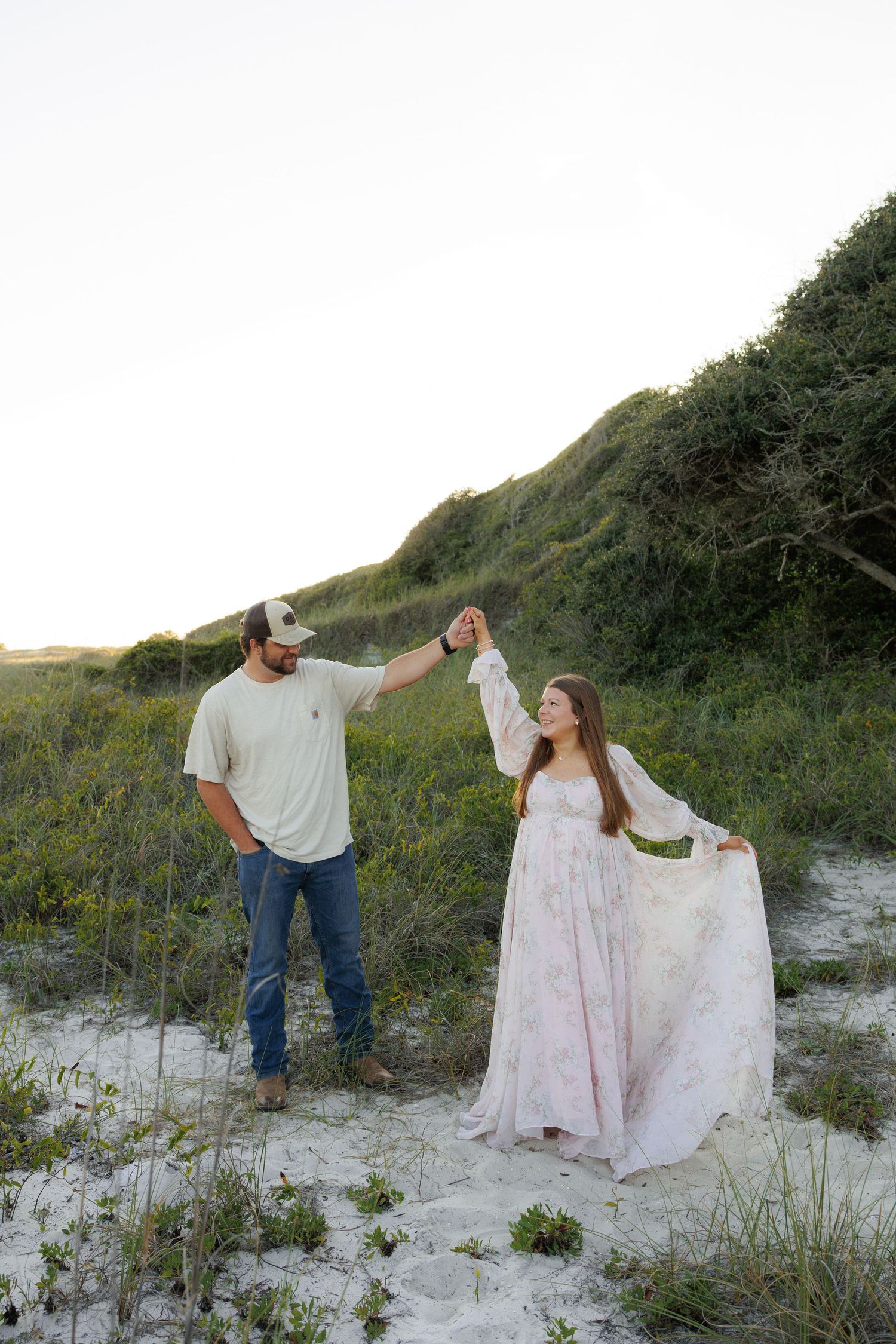 Couple dancing on sandy dunes, man in jeans and cap twirling woman in flowy floral dress, green hills backdrop.