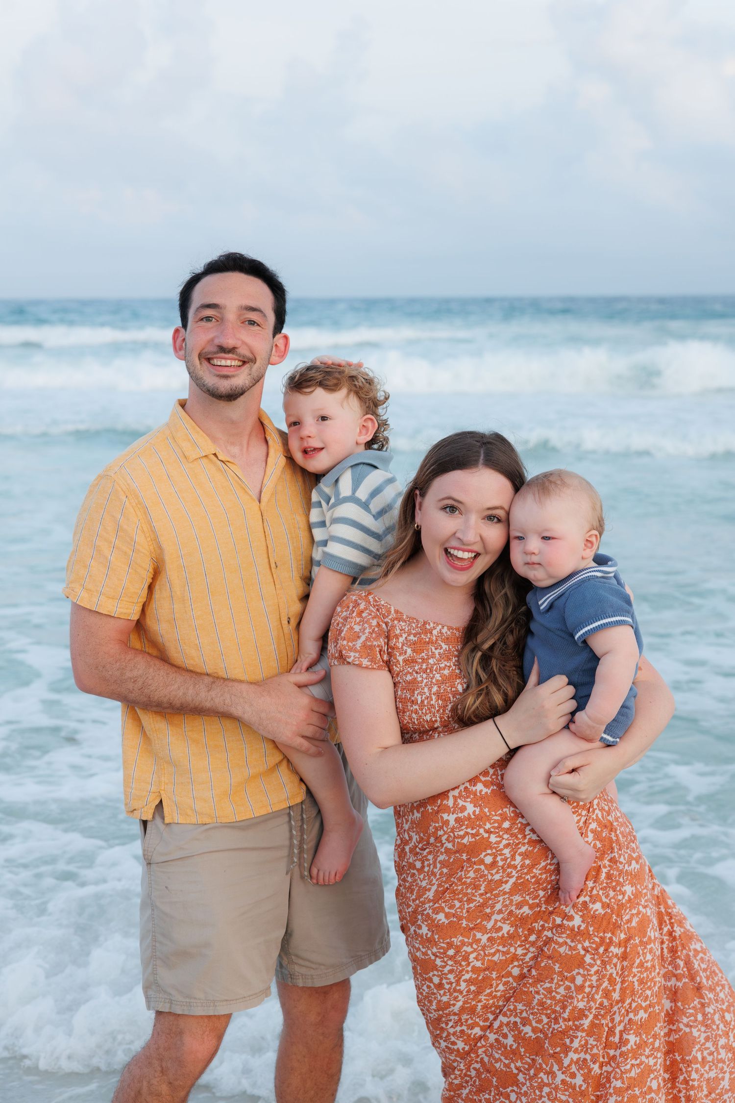 Young family with two toddler boys smiling together on a beach with ocean waves behind them.