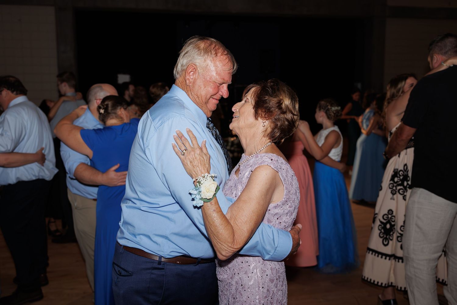 Elderly couple sharing an intimate slow dance together at a wedding reception with other dancers visible in background.
