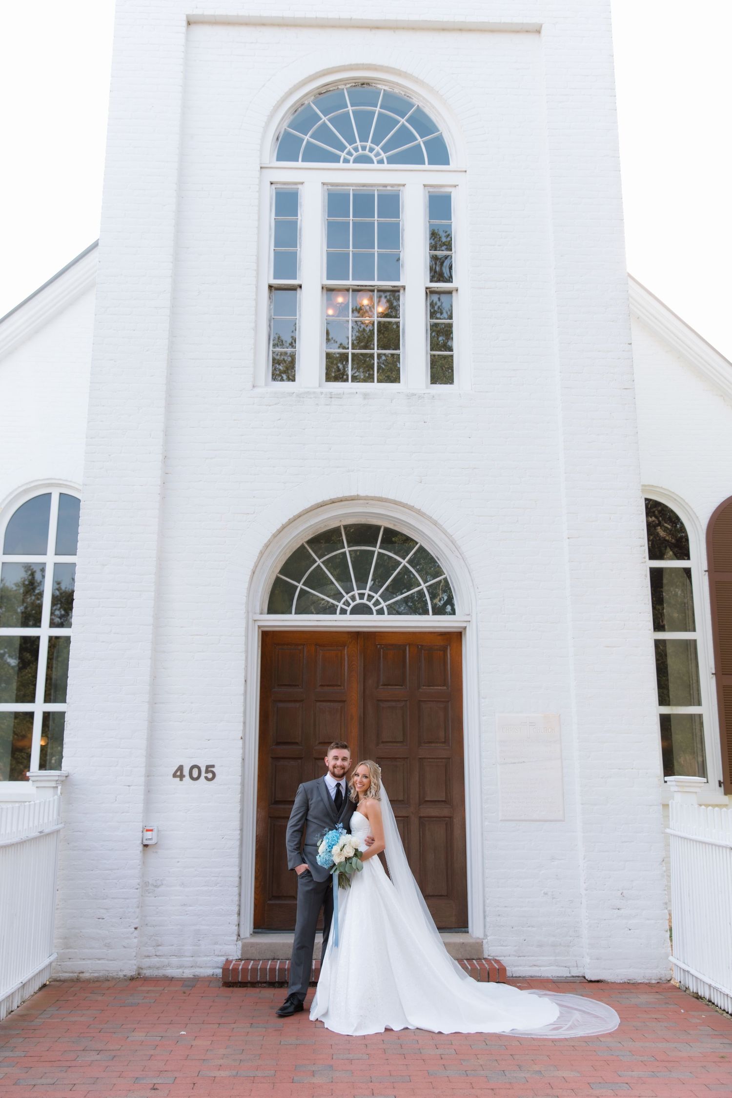 Newlyweds pose in front of classic white church with arched windows and wooden doors.