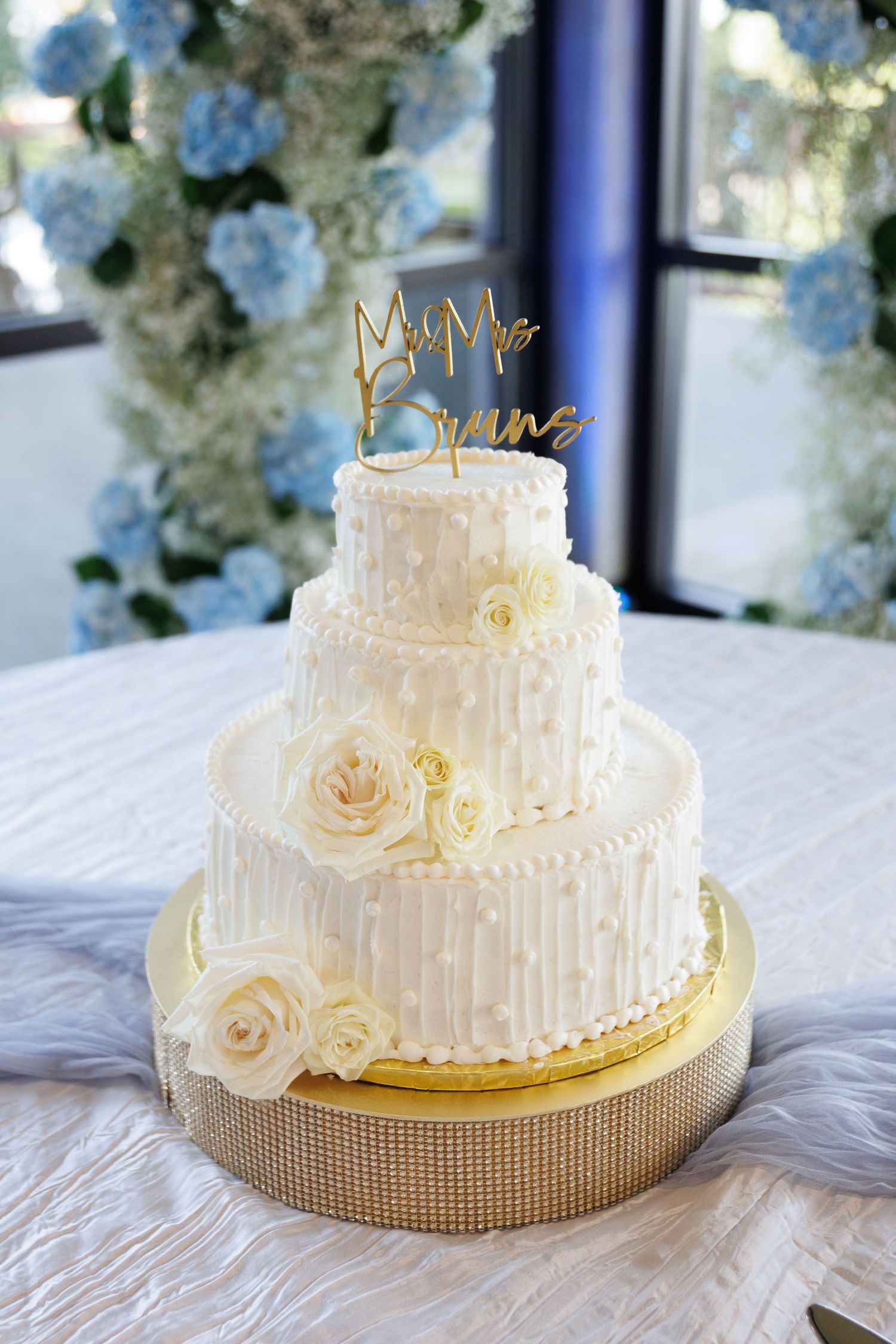 Elegant three-tier white wedding cake with roses and gold cake topper on textured stand.