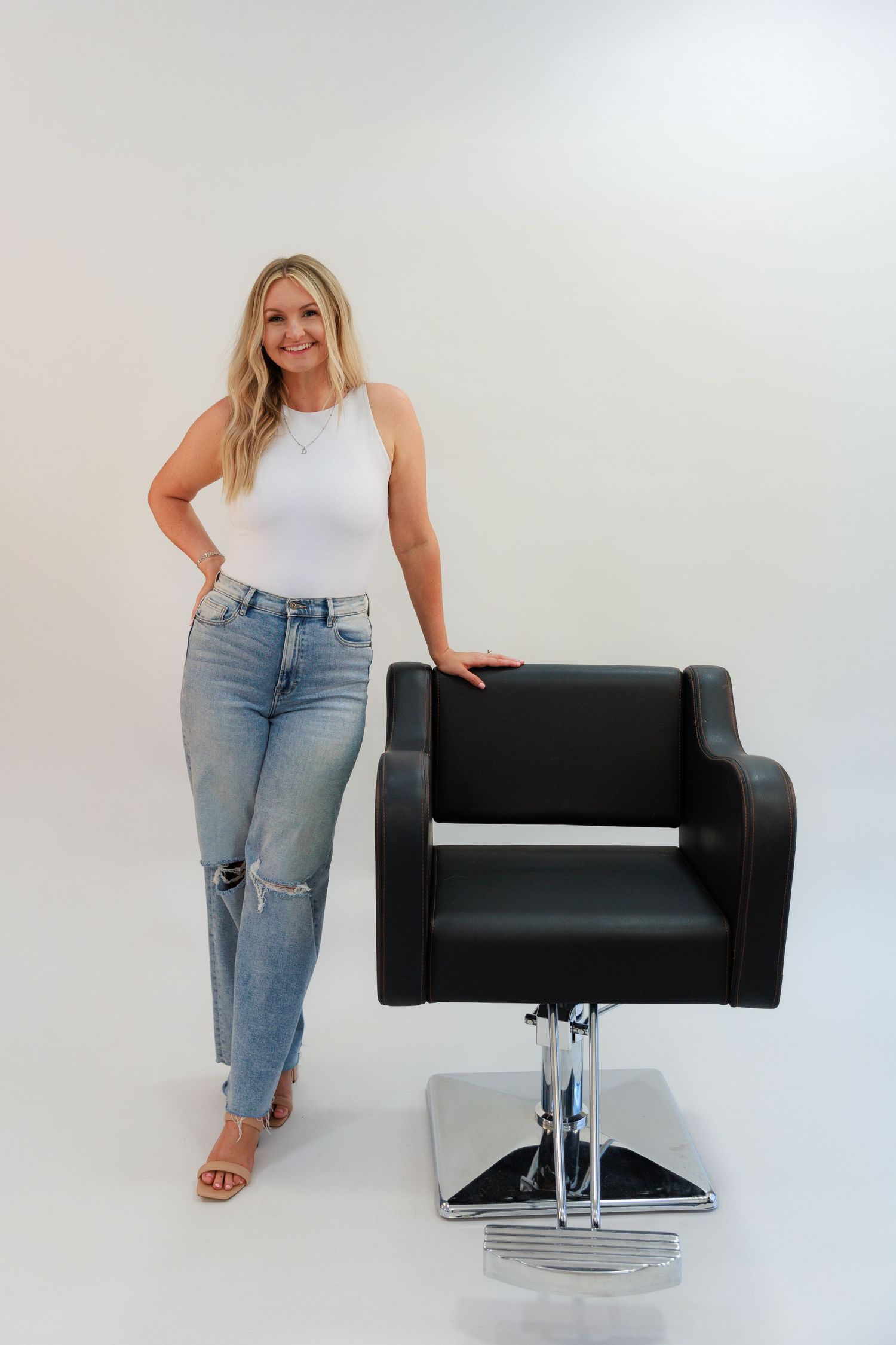 Woman in white sleeveless top and distressed jeans standing next to black salon chair.