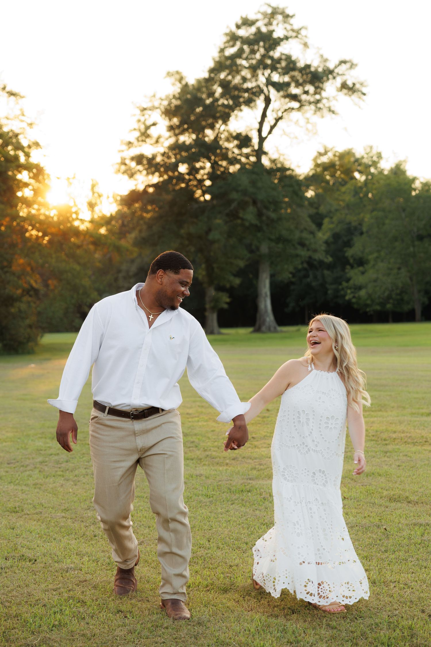 Couple holding hands and laughing while walking through a sunny green field at golden hour, wearing white and khaki.