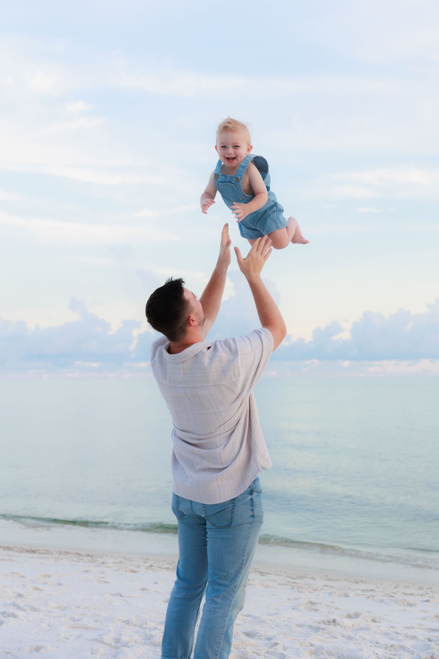 A father joyfully tosses his laughing baby into the air on a serene white sand beach at sunset.