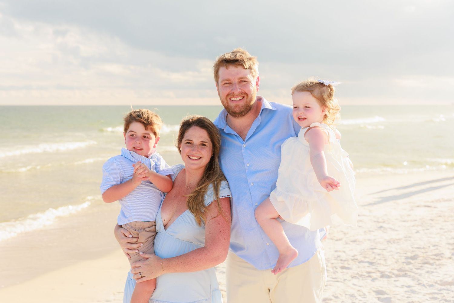 Family of four smiling on a sunny beach, parents holding two young children, with ocean waves in the background.