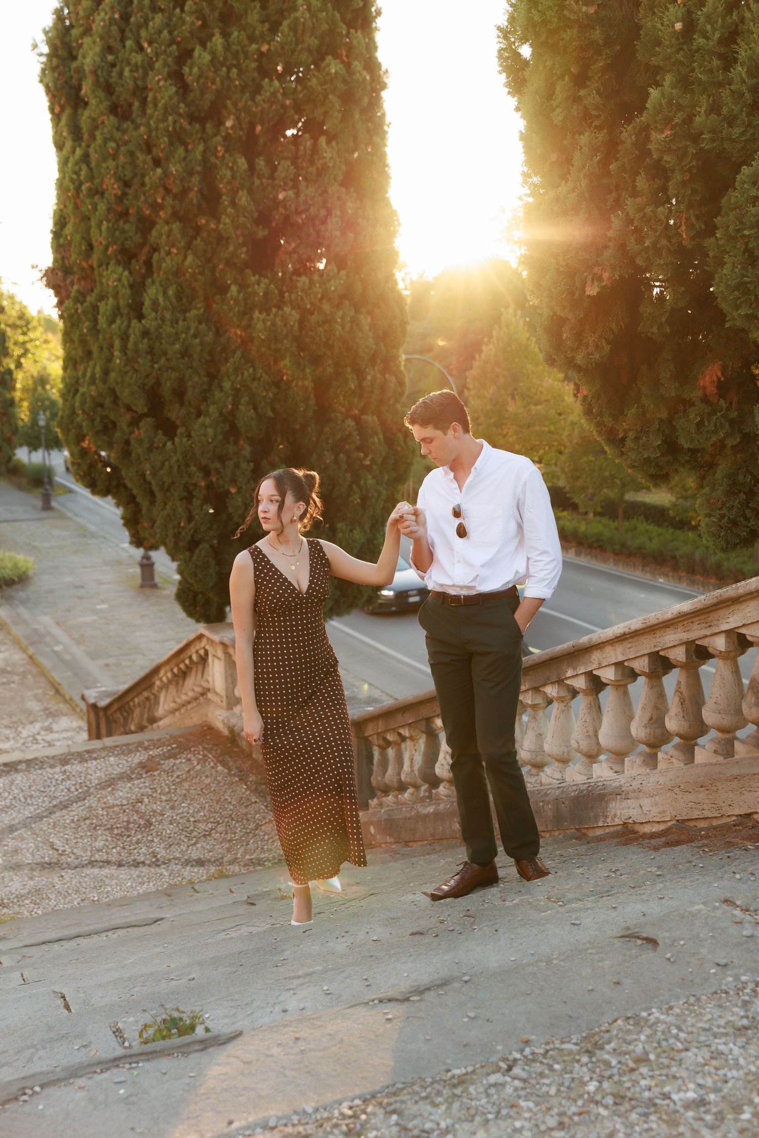 Couple walking hand in hand on stone steps at golden sunset with tall cypress trees in background.