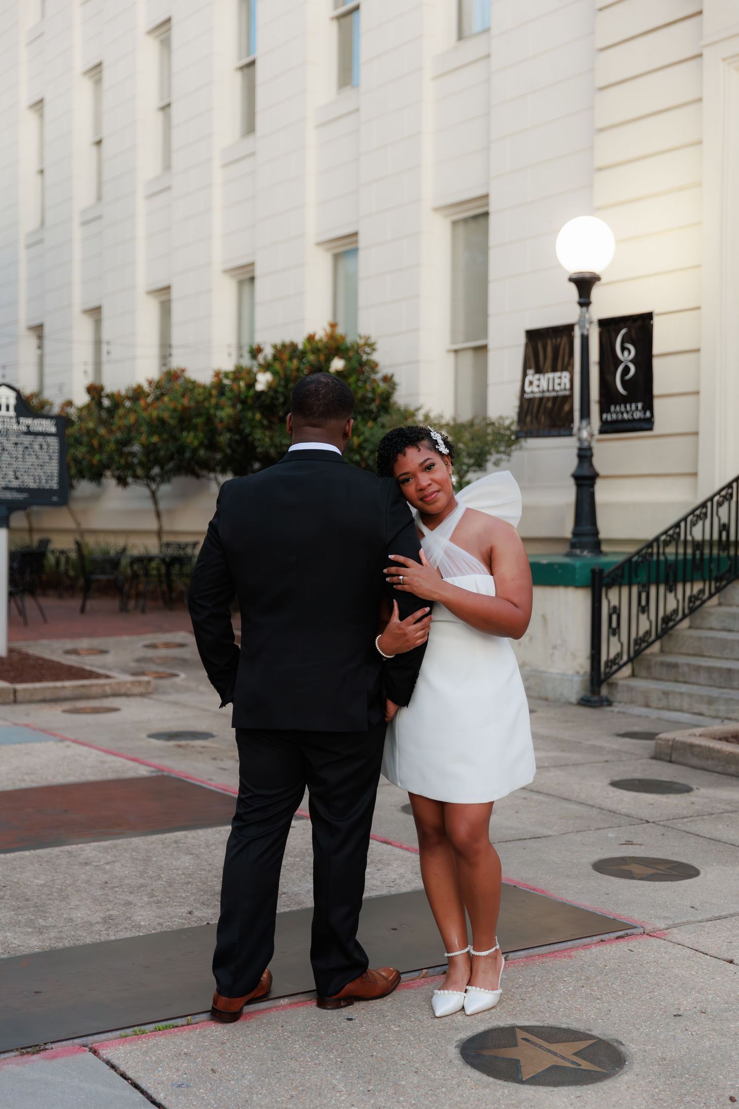 A bride in a short white halter dress and veil embraces her groom in a black suit outside a white building.