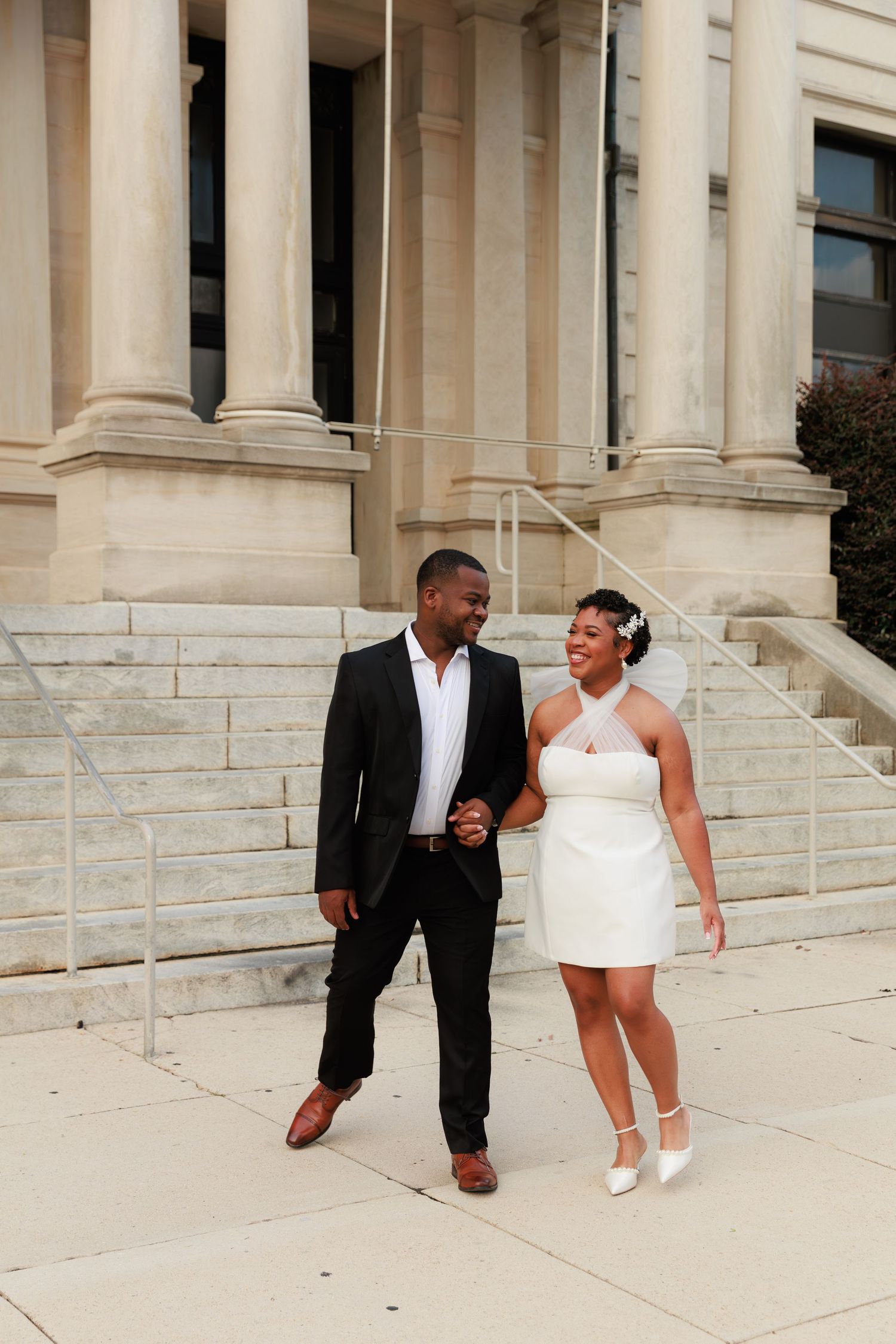 A newlywed couple holds hands and smiles while walking in front of a classic stone building with columns.