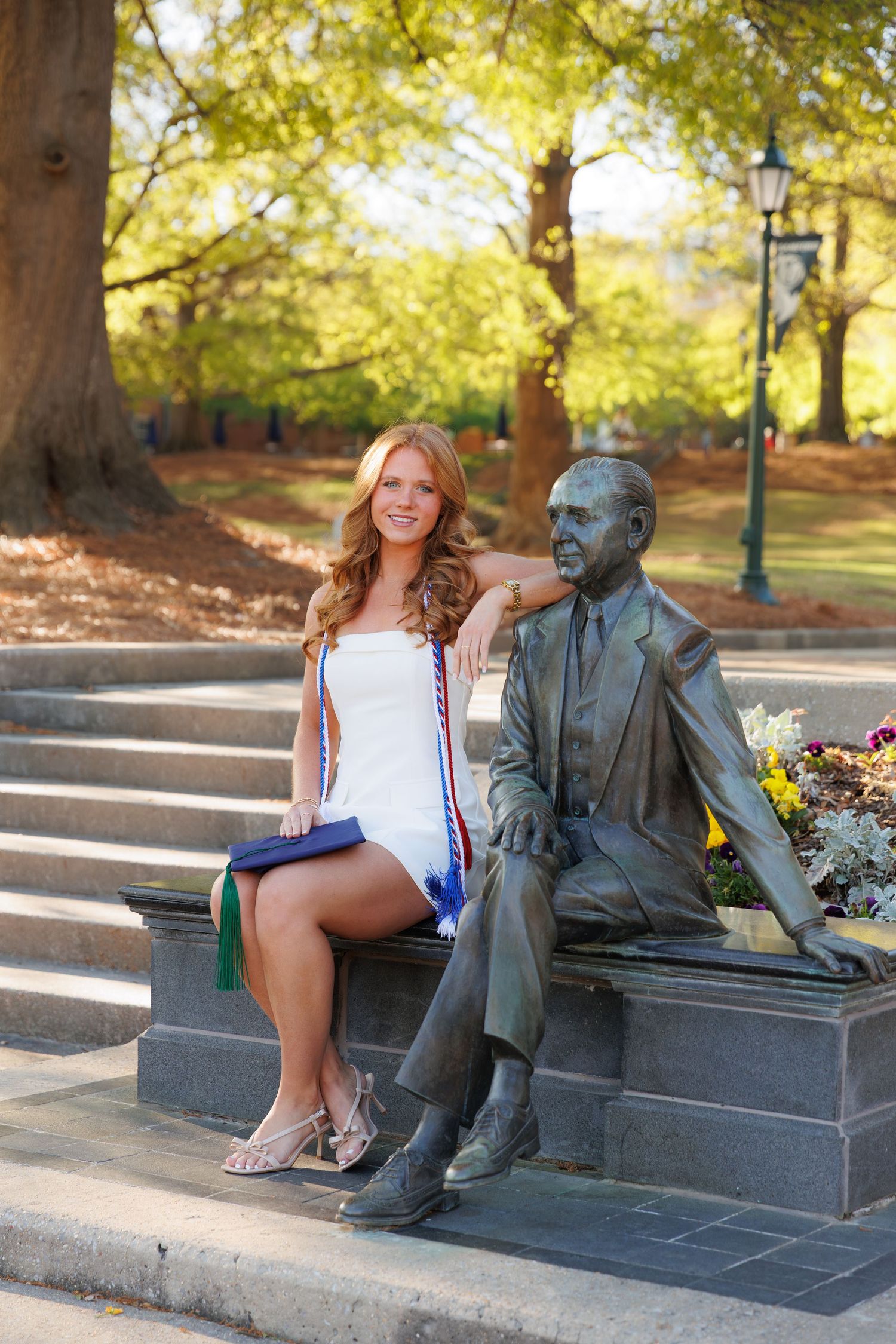 Smiling female graduate in white dress with honor cords sits beside a bronze statue on campus steps.