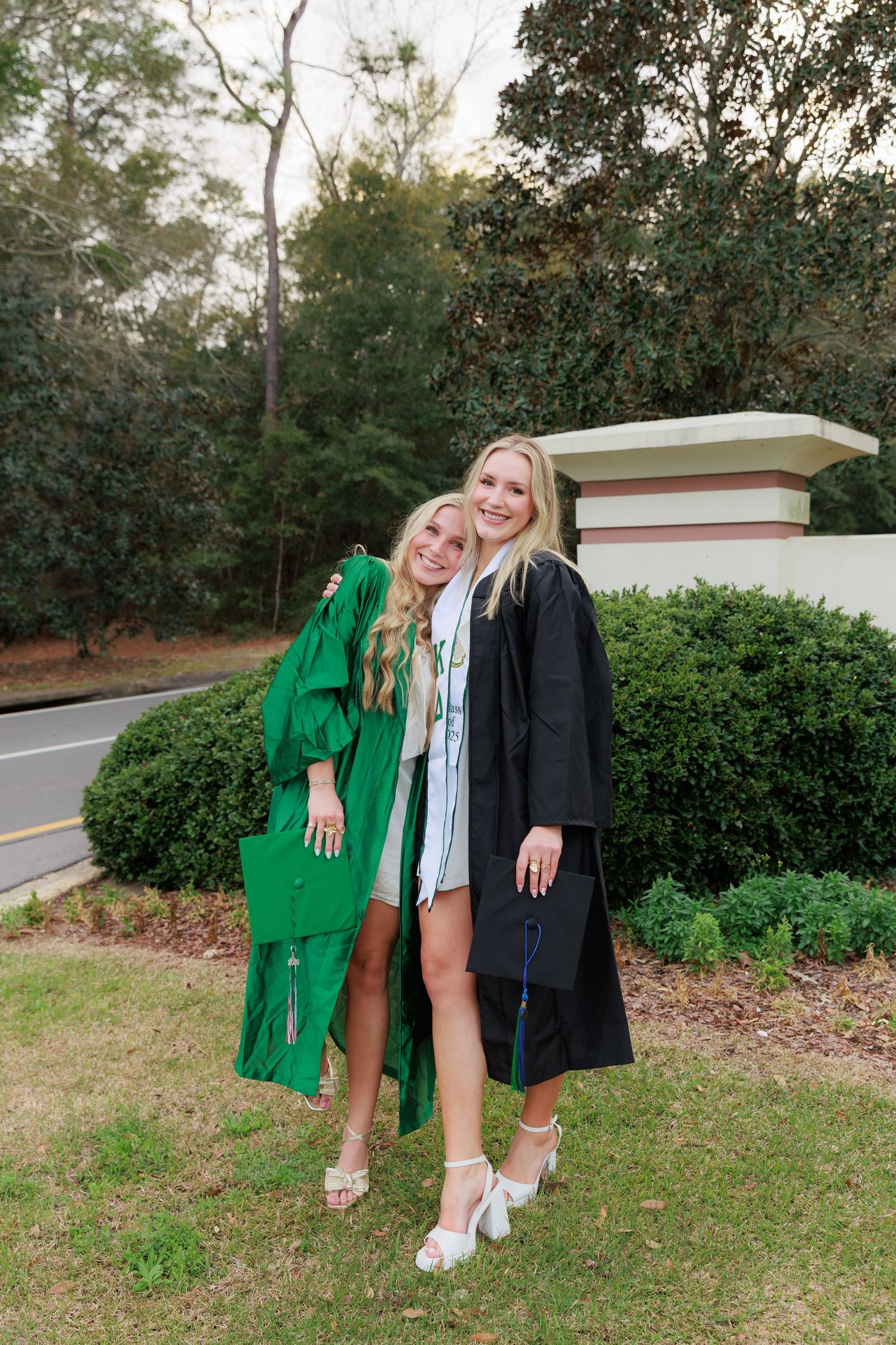Two blonde female graduates in green and black graduation gowns hugging and smiling outdoors.