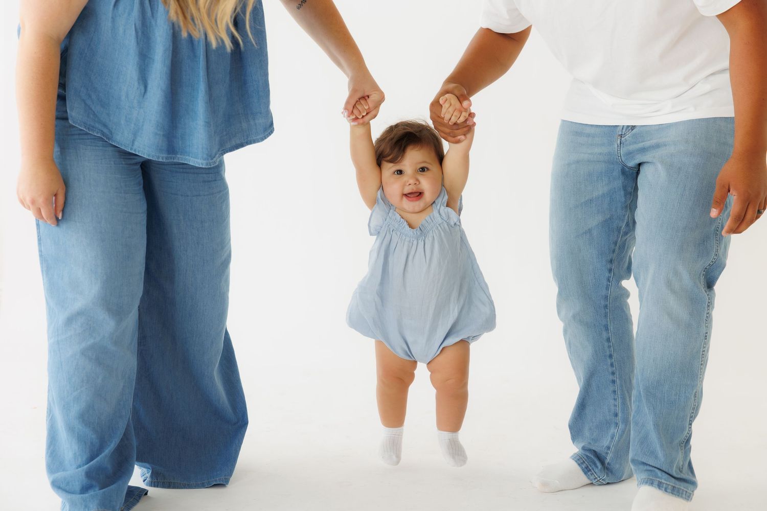 Two parents holding hands of a happy toddler girl in blue romper learning to walk on white background.