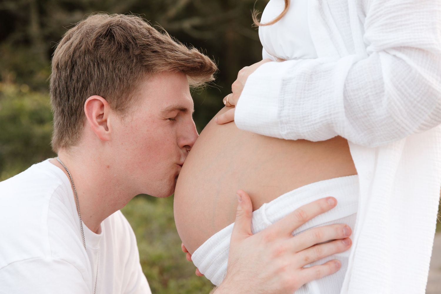 A tender moment with a person kissing a pregnant belly, both wearing white clothing outdoors.