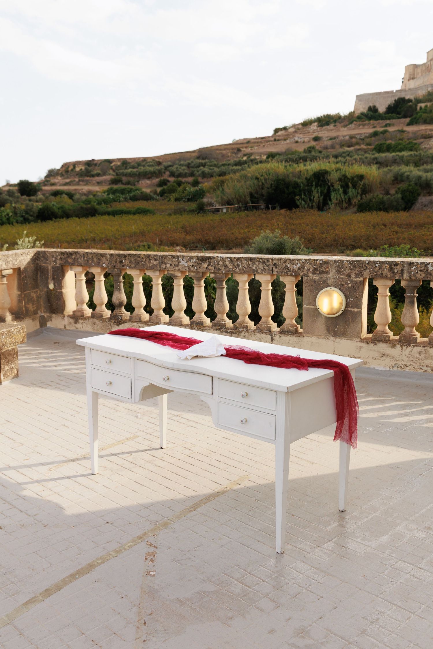 Elegant white dressing table with red fabric accent displayed on a stone terrace with ornate balustrade railing.