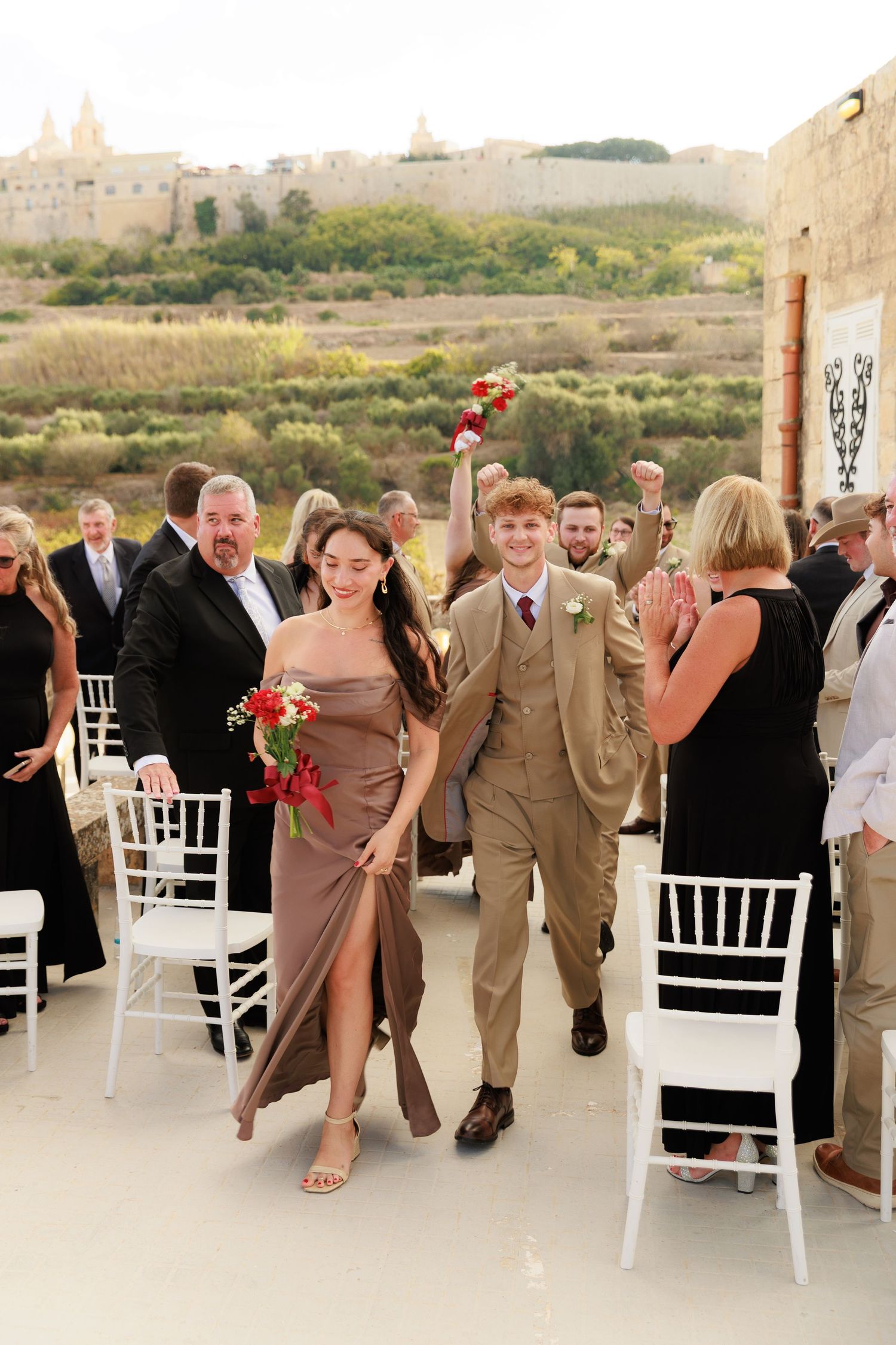 Wedding party members in tan suits dance in a line on an outdoor terrace during a celebration.