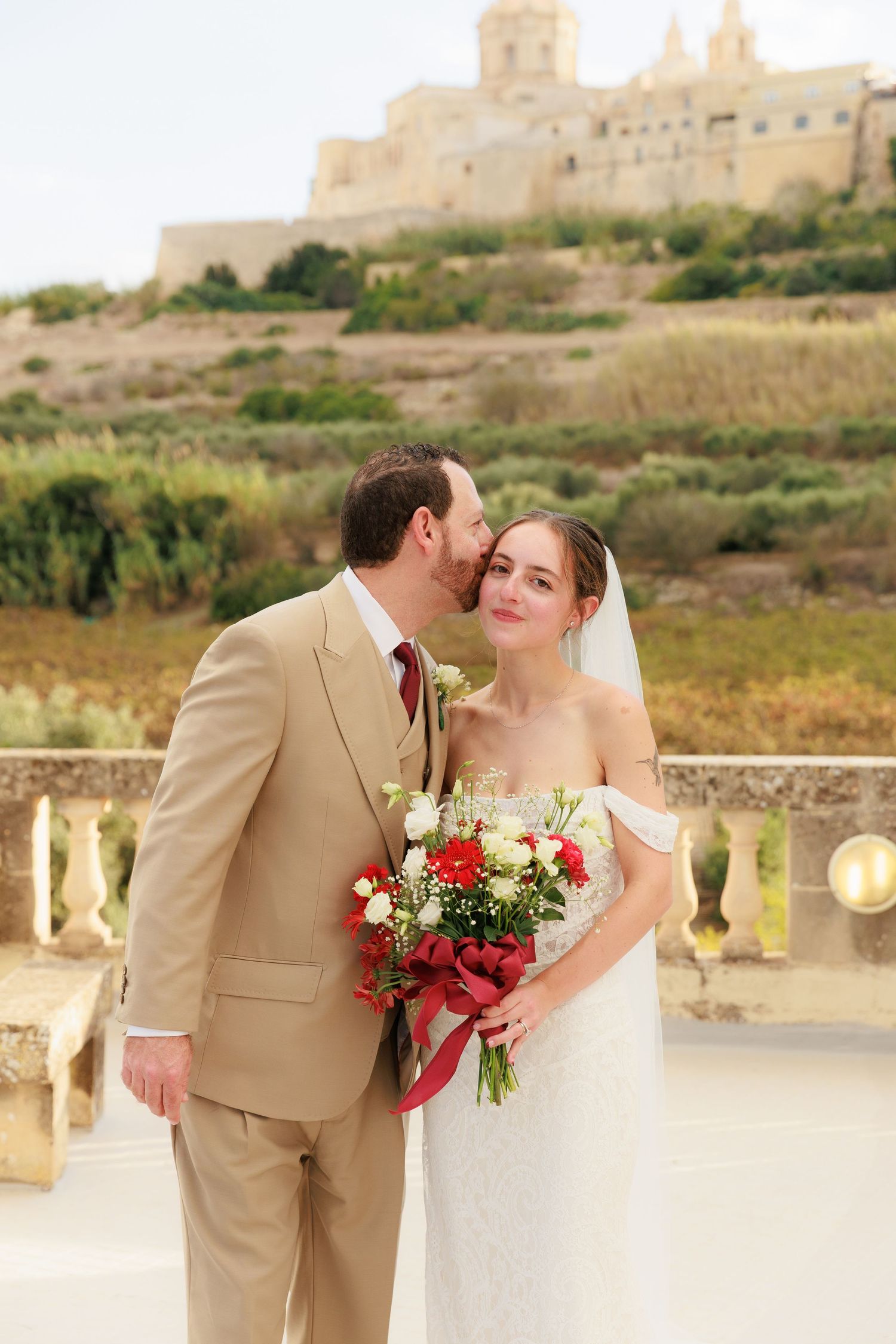 A romantic wedding couple in beige suit and white dress share a kiss against a scenic outdoor backdrop.