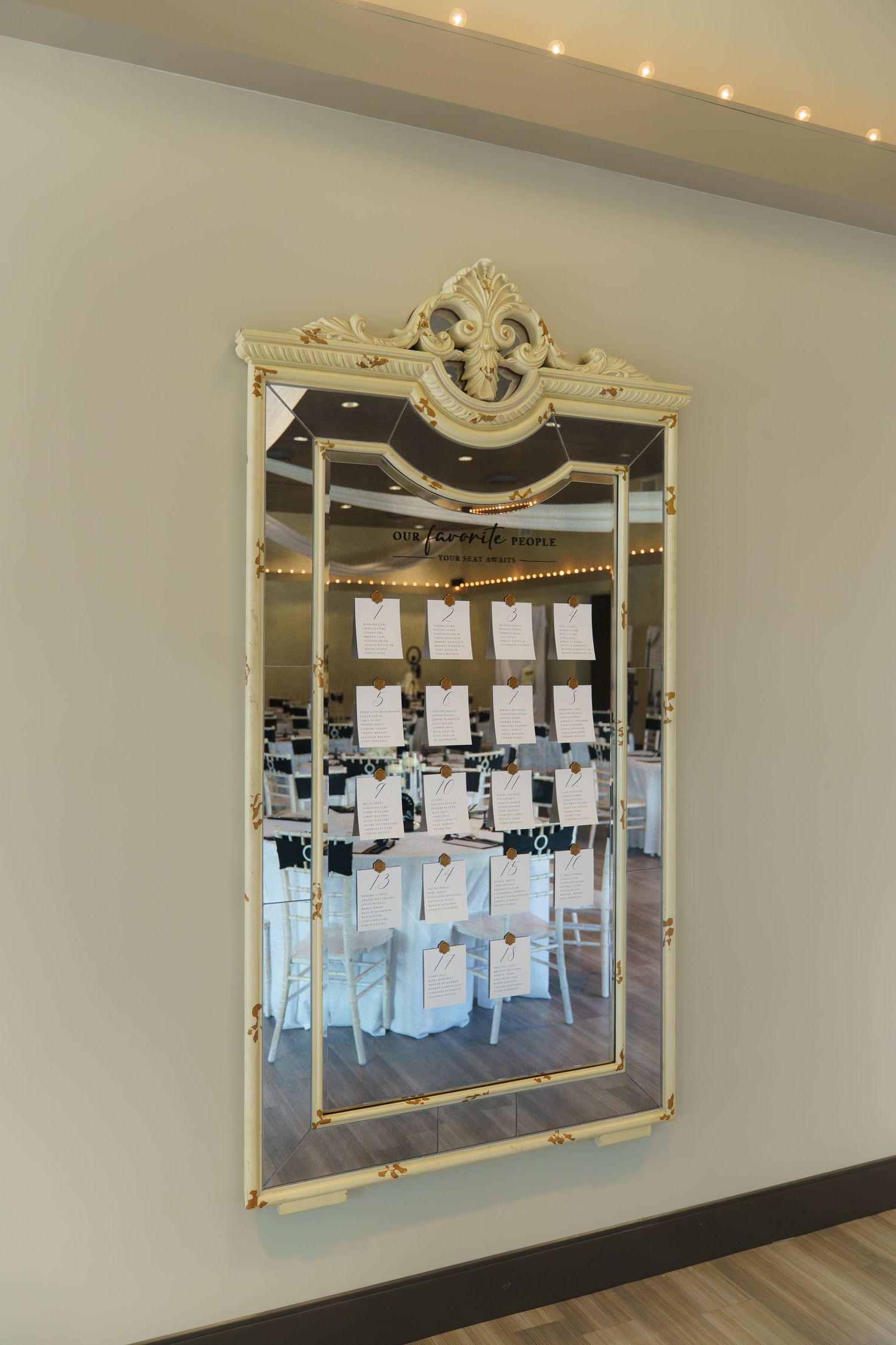An ornate gold-framed mirror displays a wedding seating chart with paper cards hanging from strings against its reflective surface.