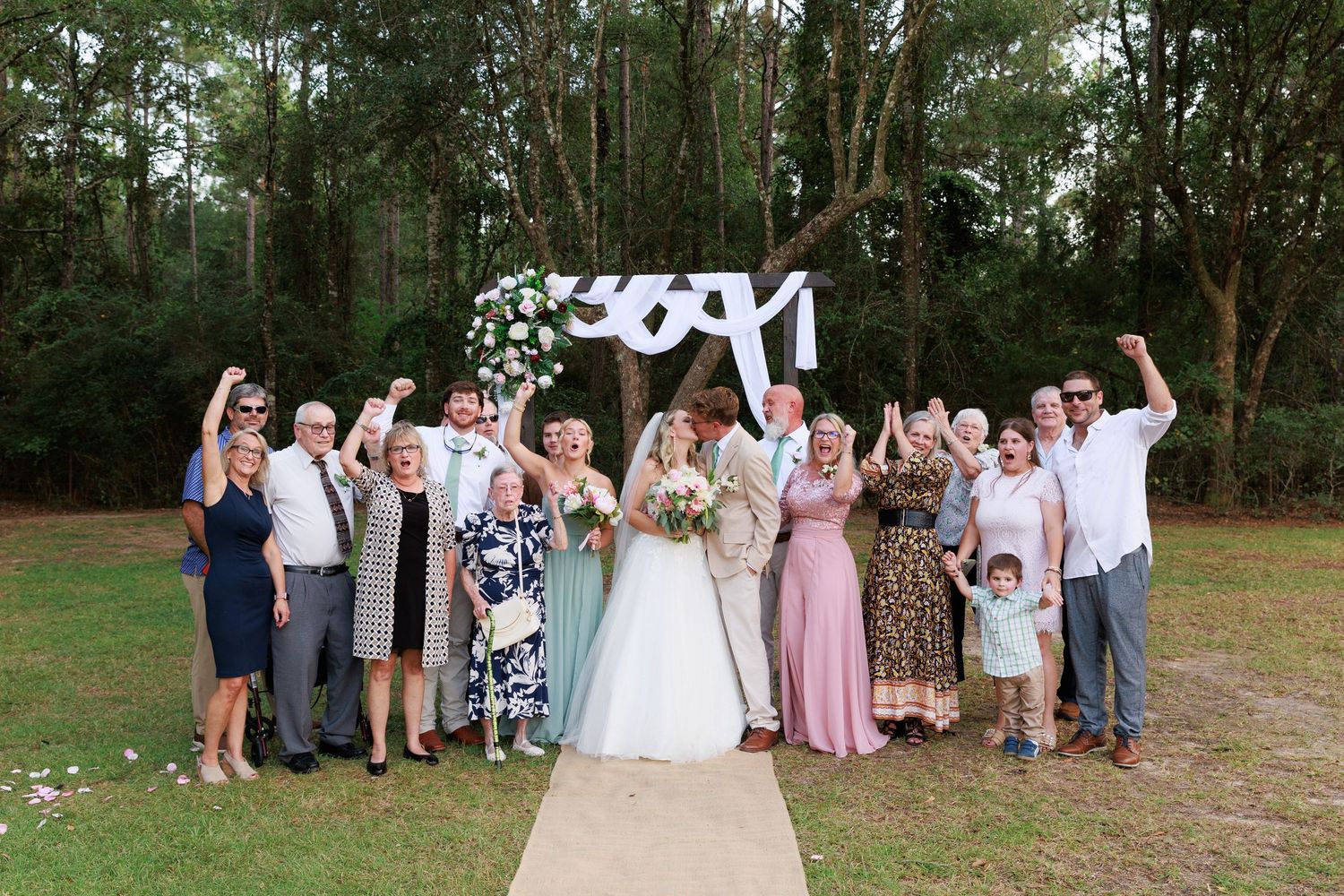 Wedding party poses together outdoors in a natural wooded setting with trees in the background.