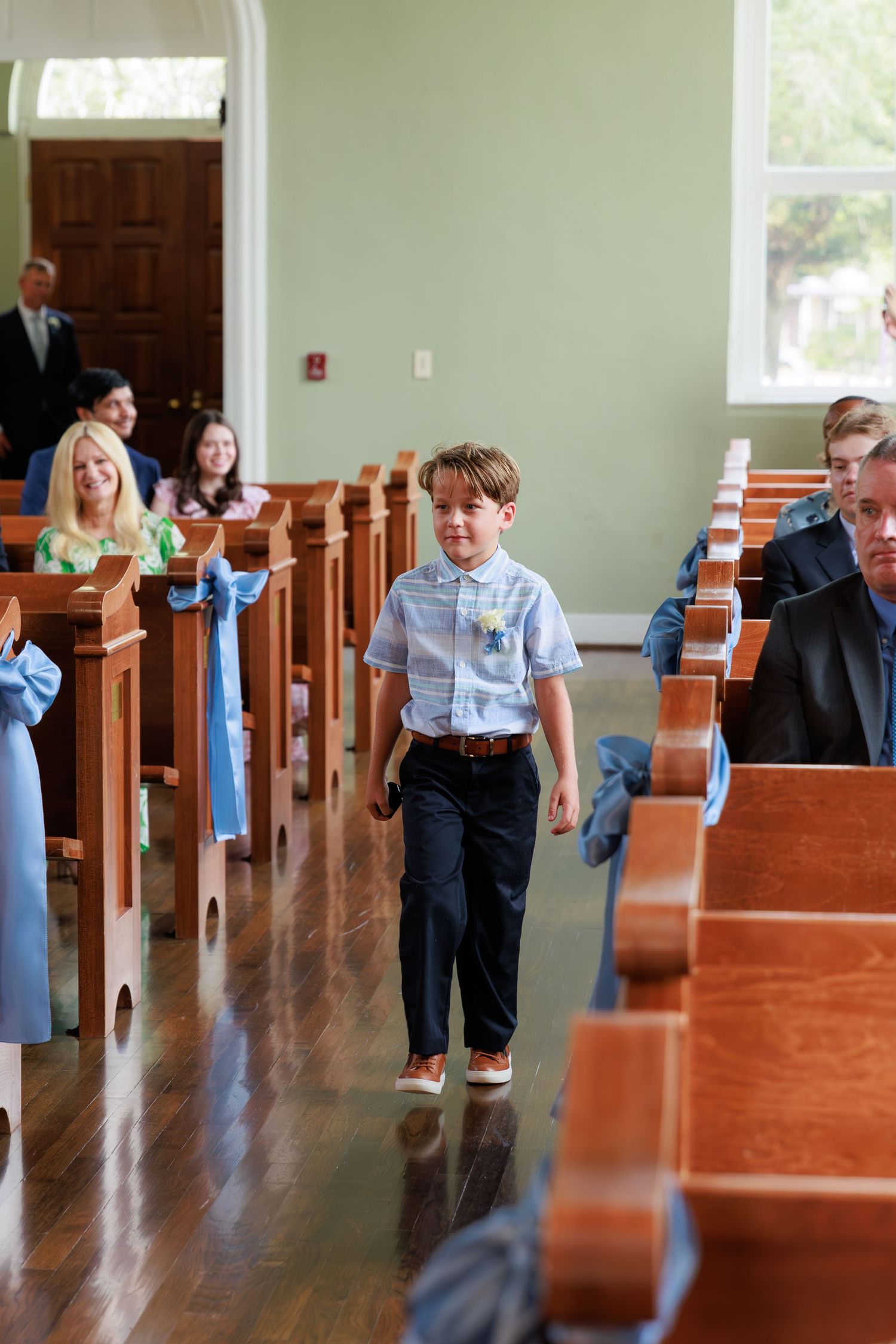 Young ring bearer in light blue shirt walks down church aisle during wedding ceremony.