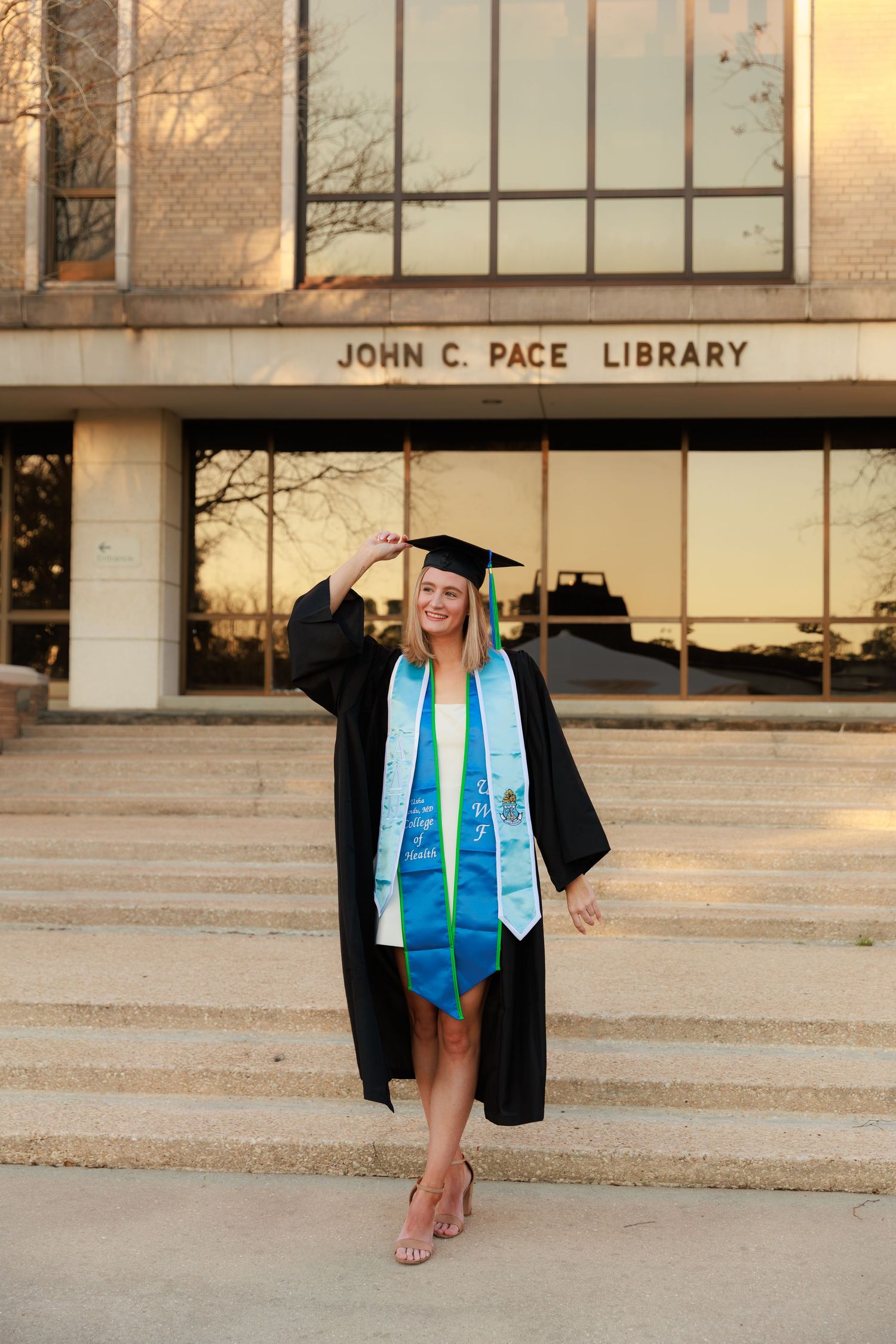 A graduate in cap and gown stands on steps outside the John C. Pace Library wearing blue graduation stoles.
