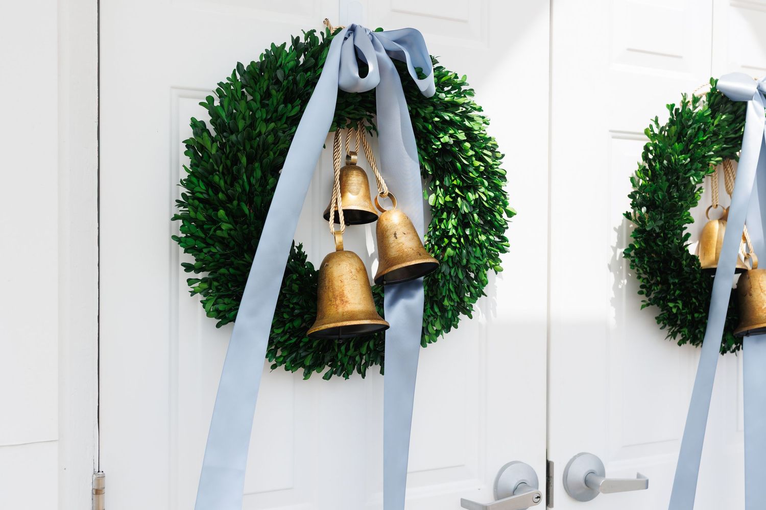 Gold bells with blue ribbon hang from a lush green boxwood wreath mounted on a white wall.
