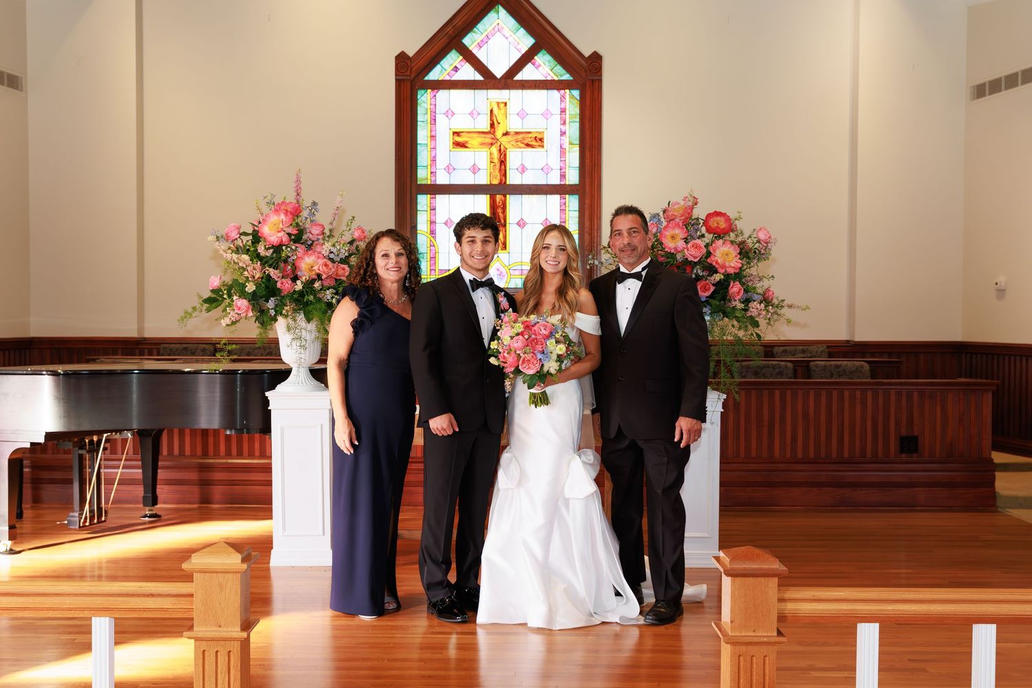 Small church wedding party stands together at altar with stained glass cross window and pink floral arrangements.