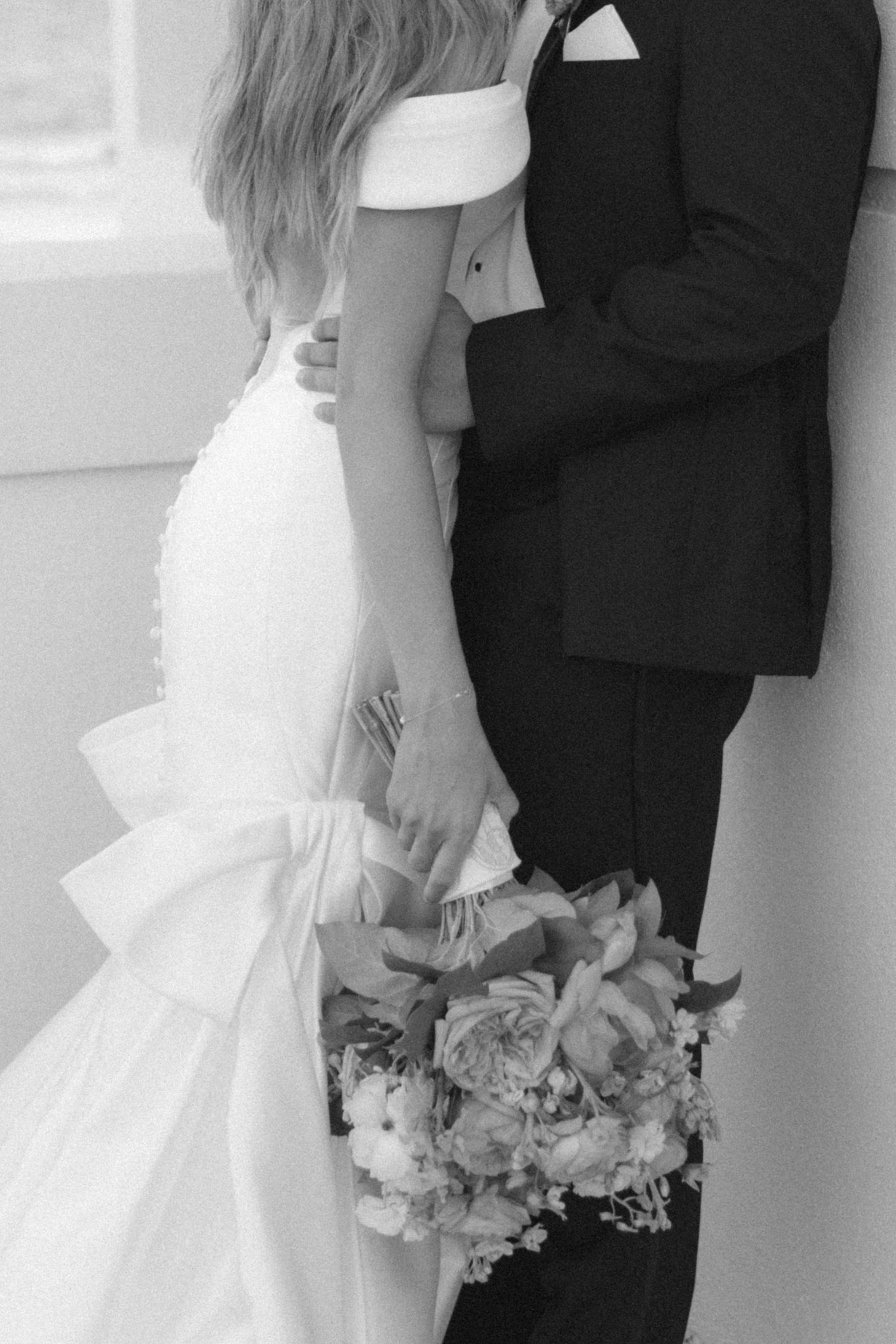 Black and white photo of couple embracing in wedding attire, with floral bouquet held between them.