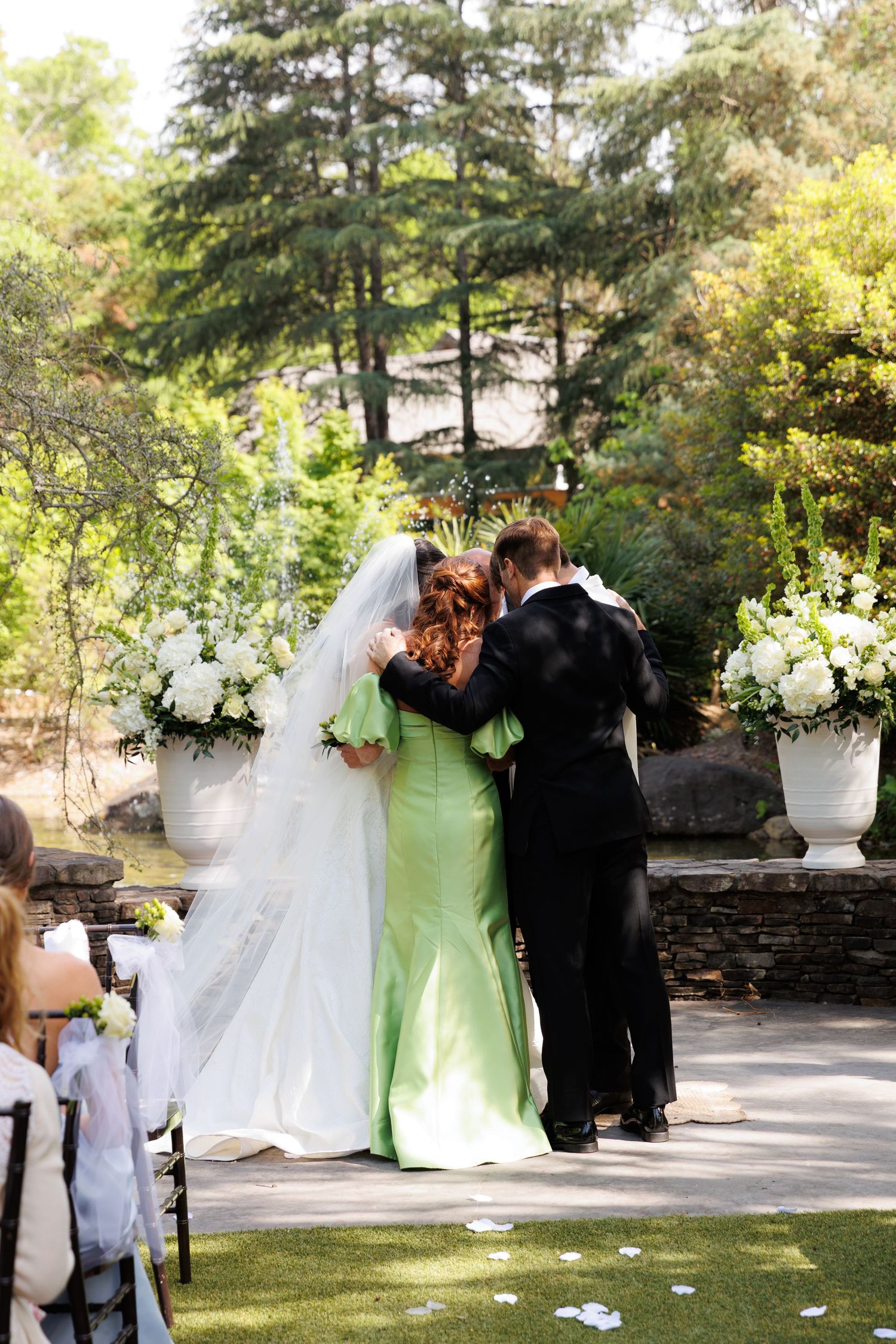 Bride in white gown embraced by woman in green dress at outdoor wedding altar with white floral arrangements.