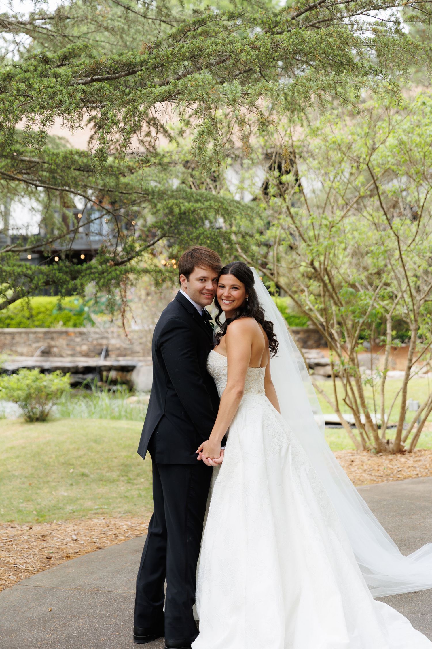 Smiling bride in ball gown and groom in tuxedo pose together in garden with veil trailing behind them.
