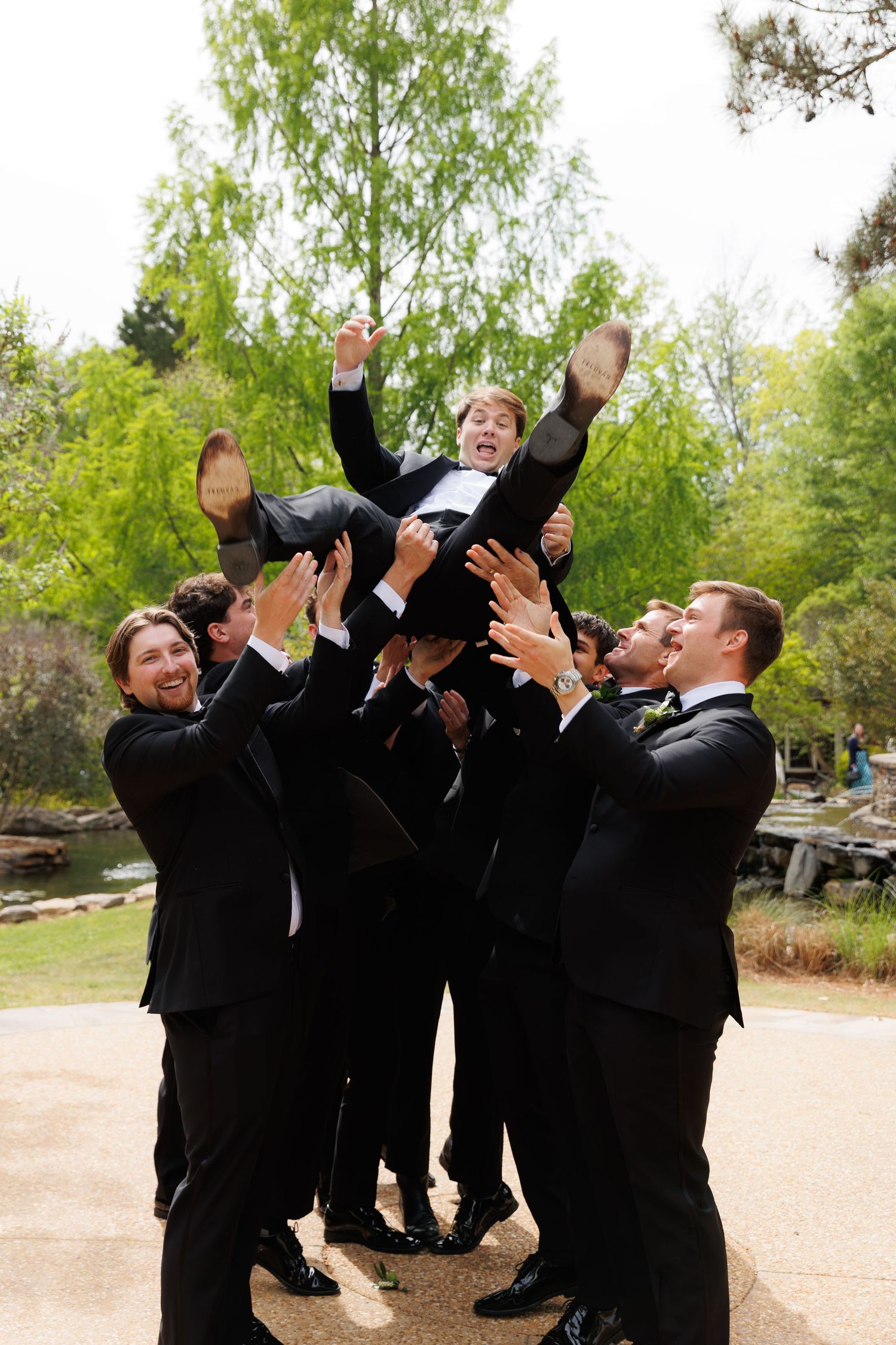 Groomsmen in black tuxedos joyfully tossing the groom into the air outdoors at a wedding celebration.