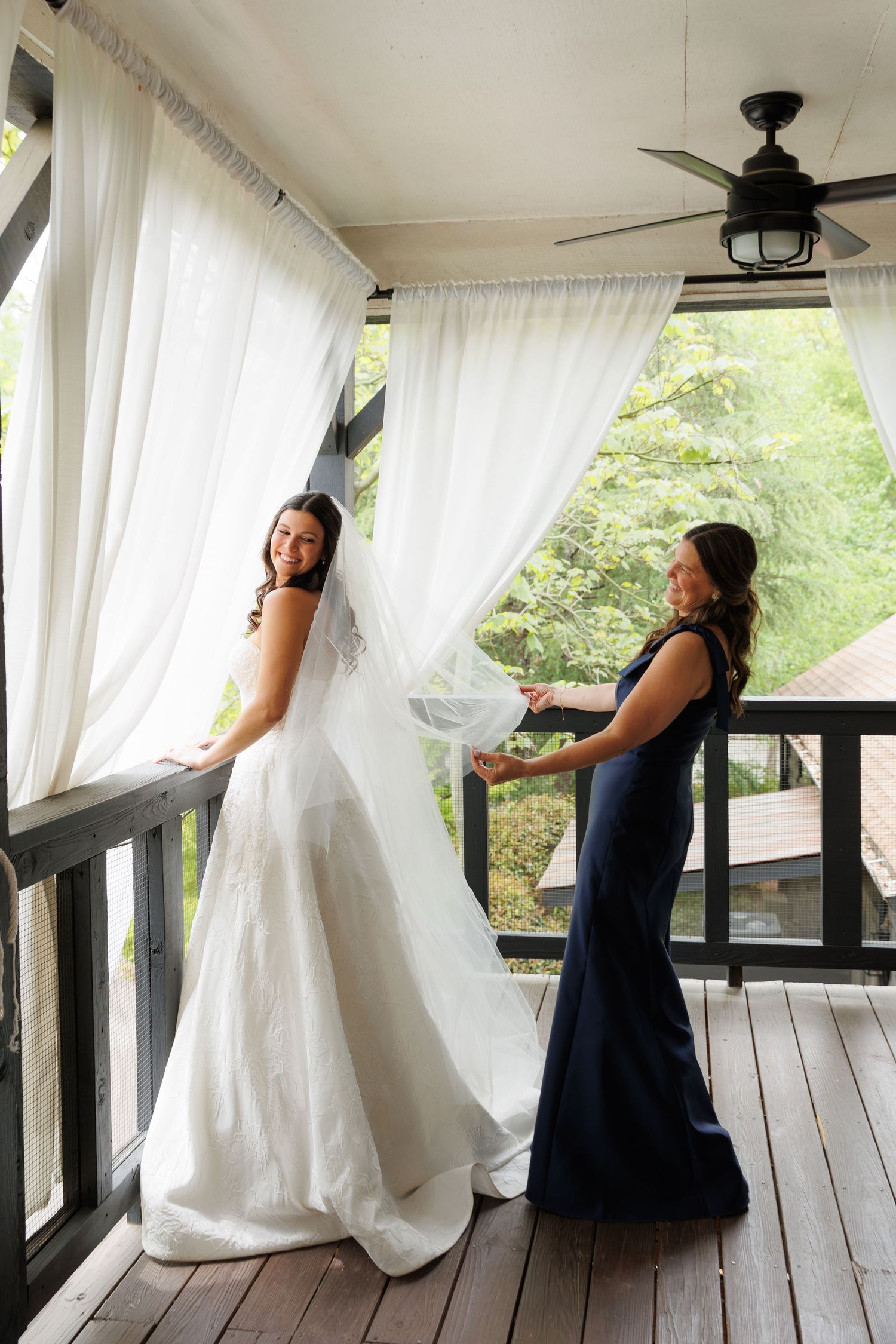 Bride in elegant white gown smiles on porch while bridesmaid in navy dress adjusts her veil.