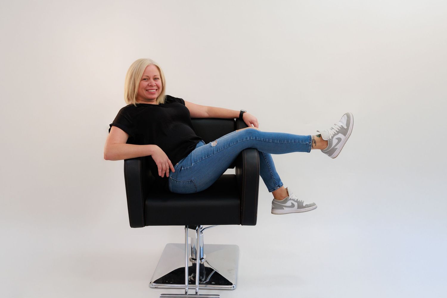 Relaxed pose in black salon chair wearing casual attire and silver sneakers.