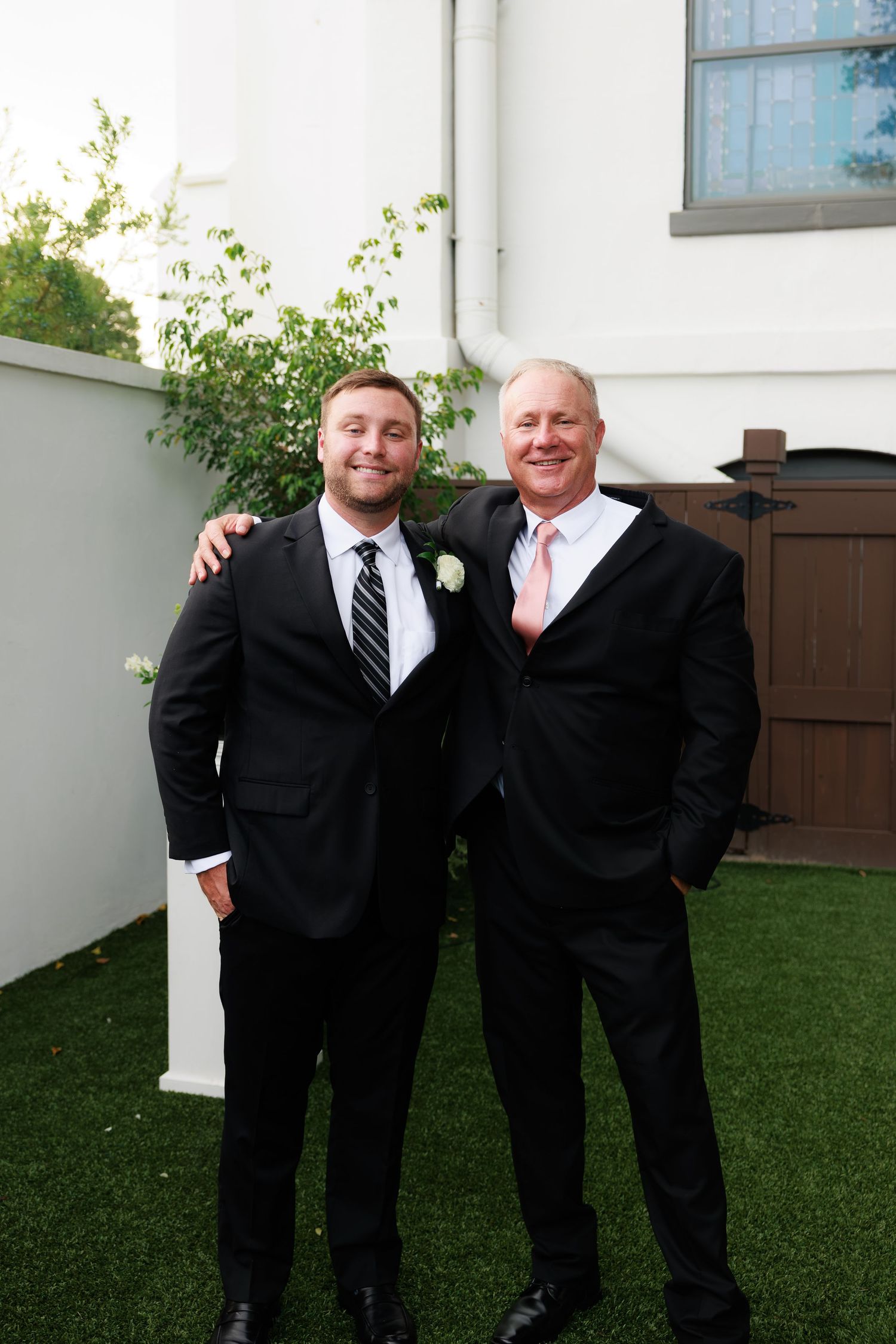 Two men in black suits smiling outdoors at a wedding, one wearing a boutonniere and striped tie.