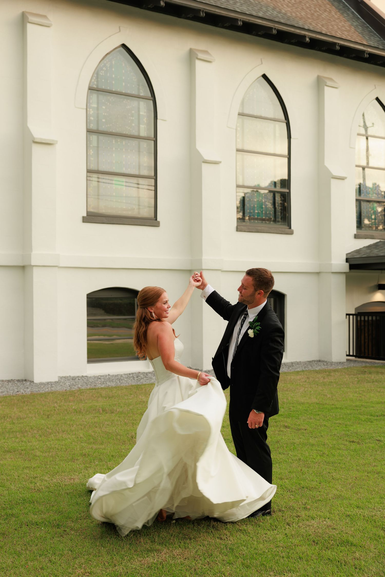 Groom twirls bride in white gown on church lawn, both laughing joyfully during outdoor wedding portraits.