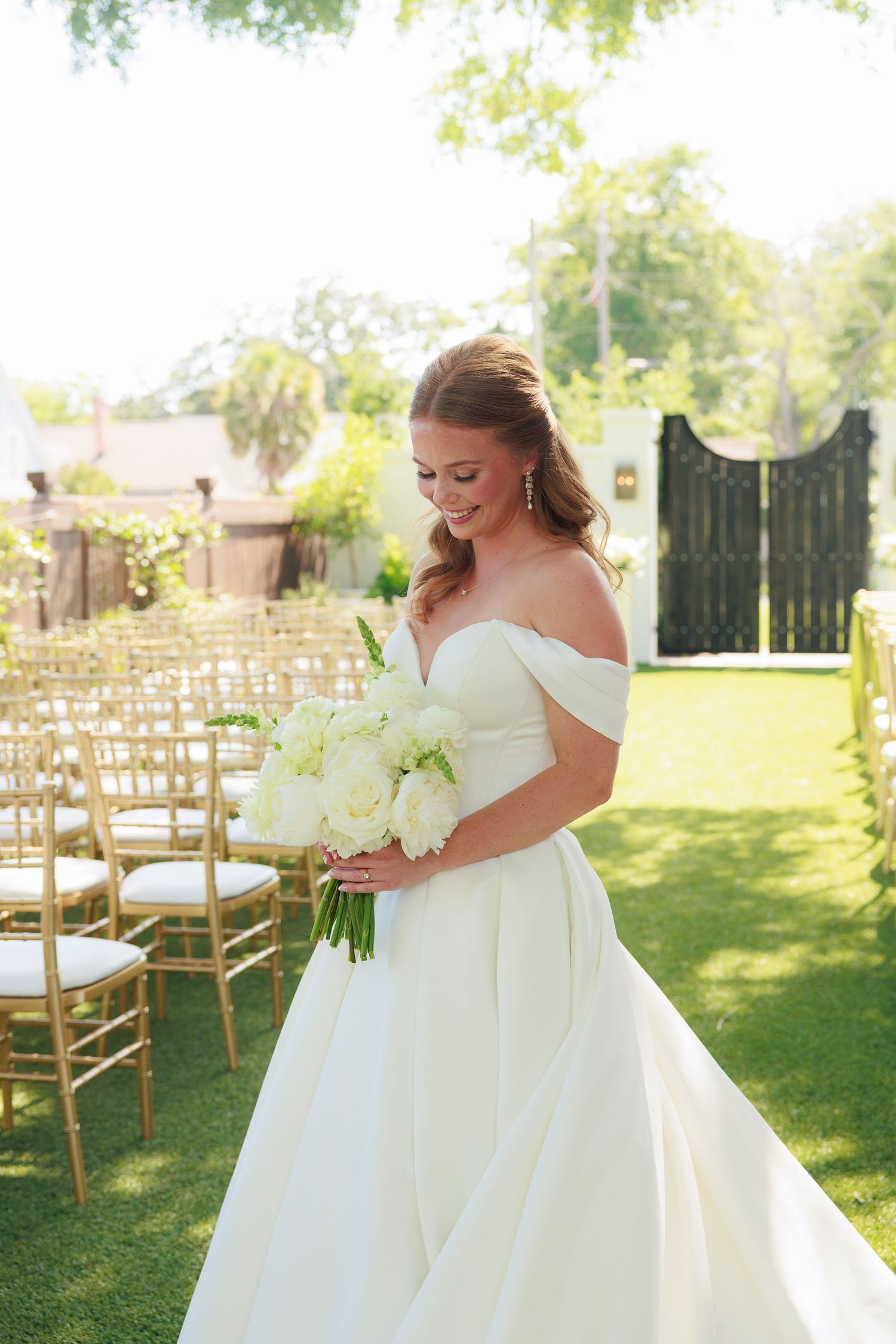 Elegant bride in off-shoulder satin gown gazing at white floral bouquet at outdoor wedding ceremony venue.