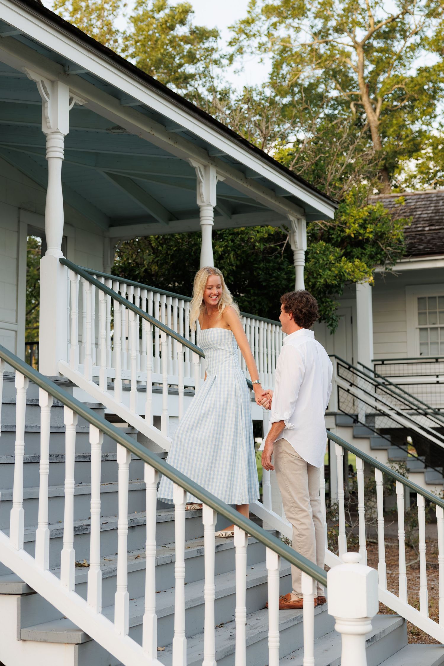 A couple holds hands on white porch stairs, woman in striped dress smiling at man in white shirt.