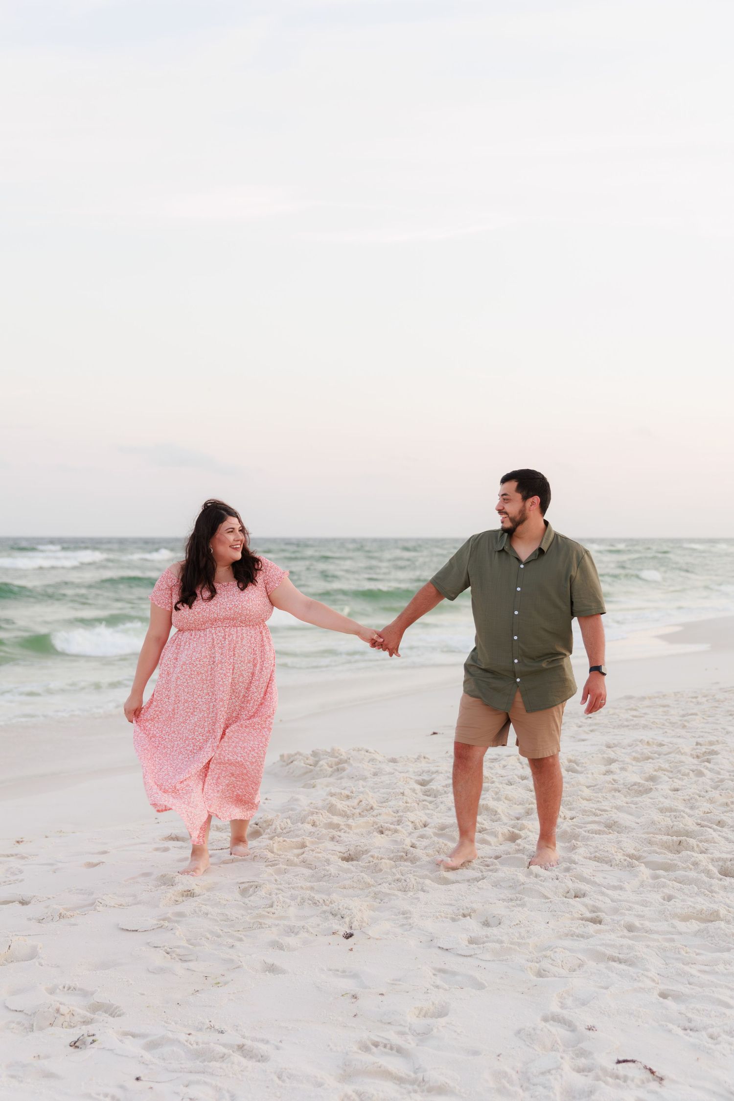 Couple holding hands walking on a white sand beach at sunset, woman in pink dress, man in green shirt.