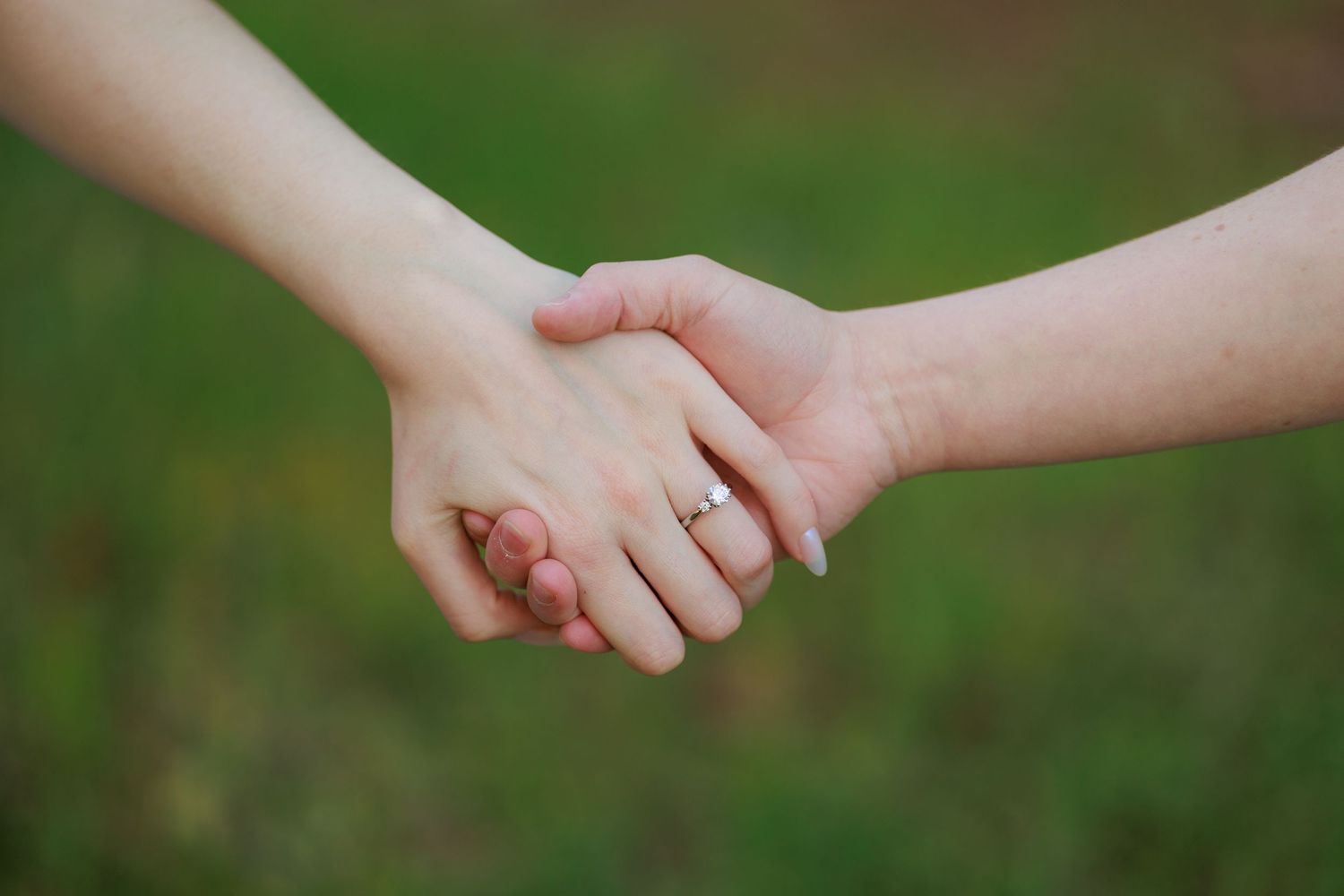 Close-up of a couple holding hands with an engagement ring visible on a blurred green background.