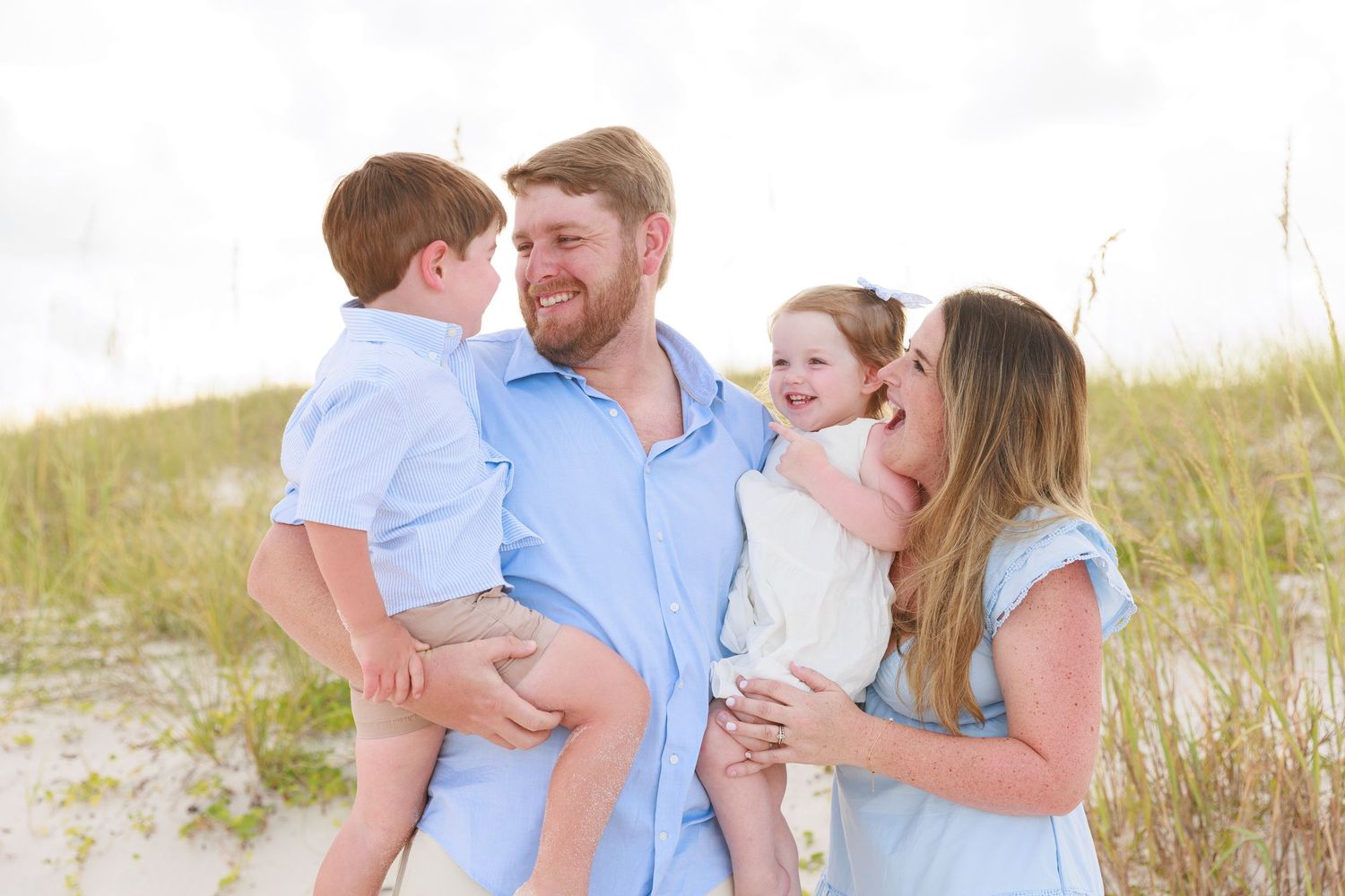 Happy family of four enjoying a sunny beach day, parents holding young children among coastal dunes and sea grass.
