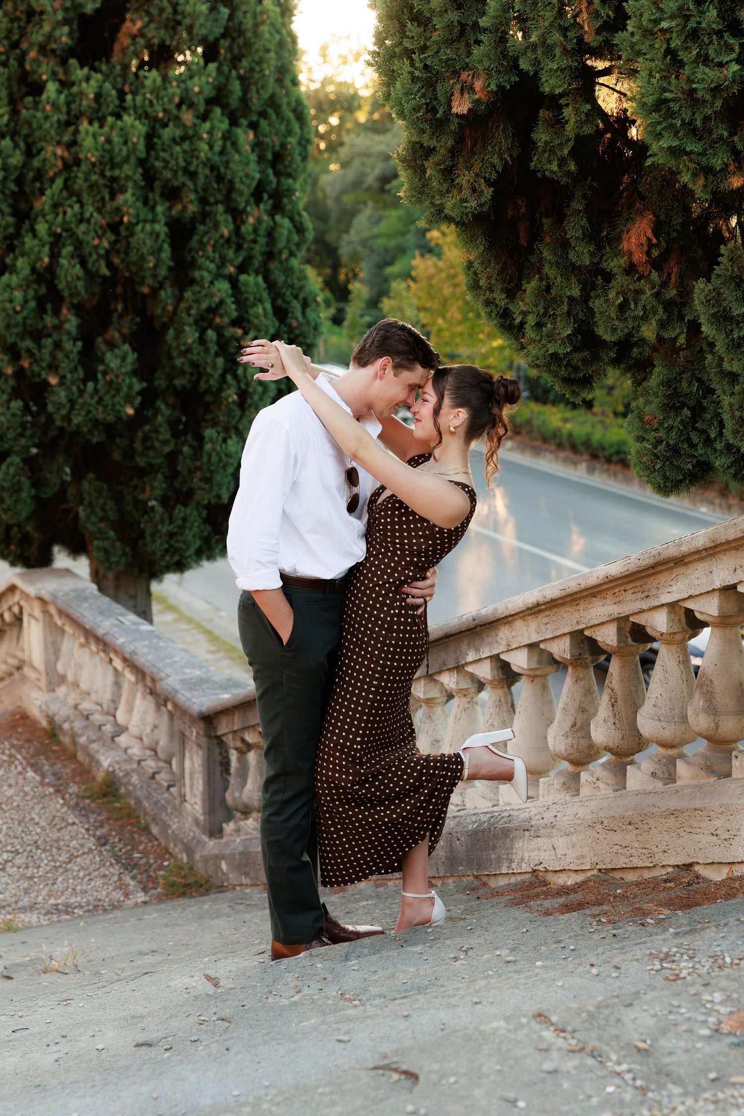 Lovers sharing romantic dip embrace on terrace steps surrounded by cypress trees at golden hour.