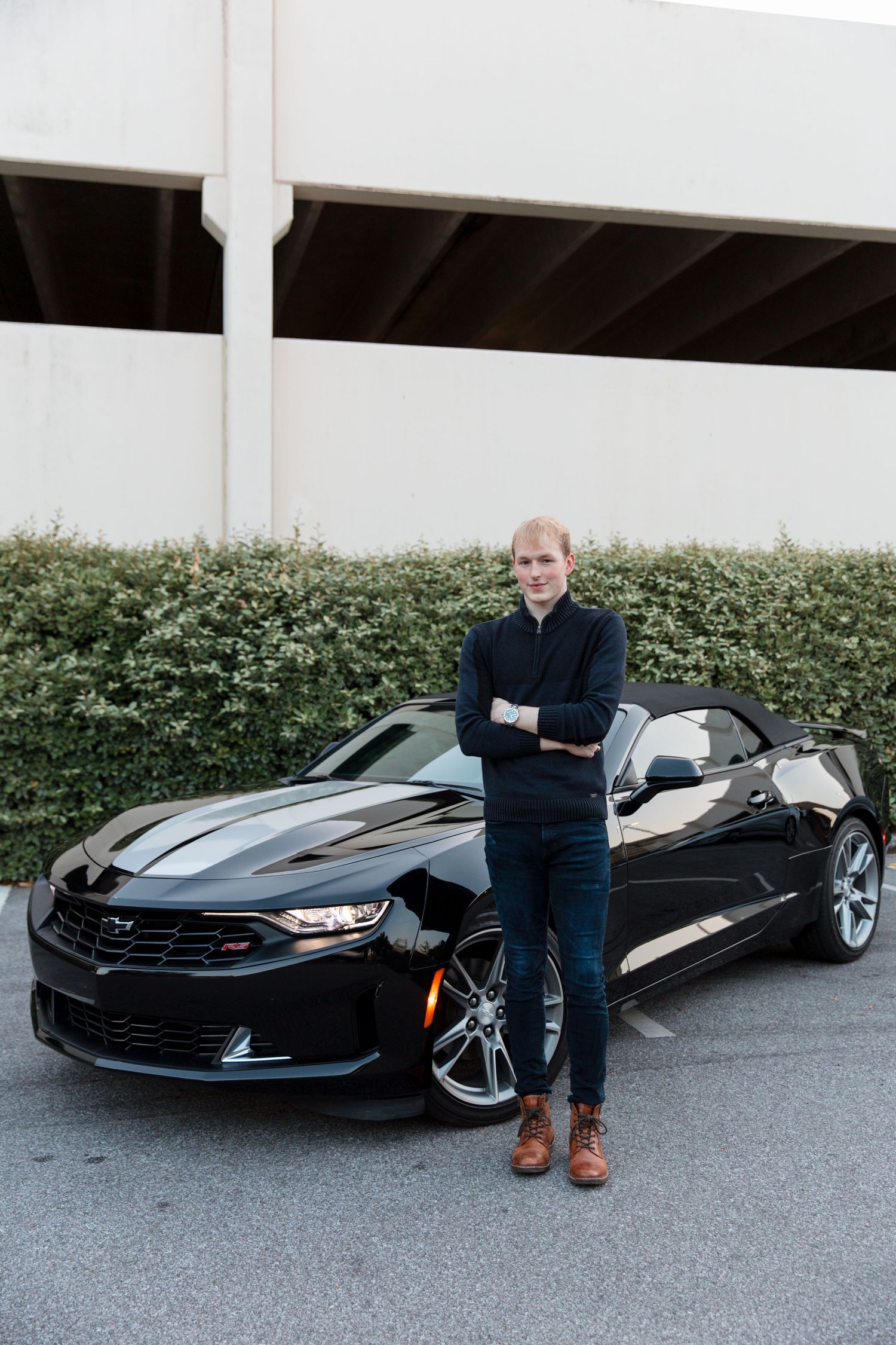 Man in dark outfit stands arms crossed beside a black Chevrolet Camaro convertible in a parking area.