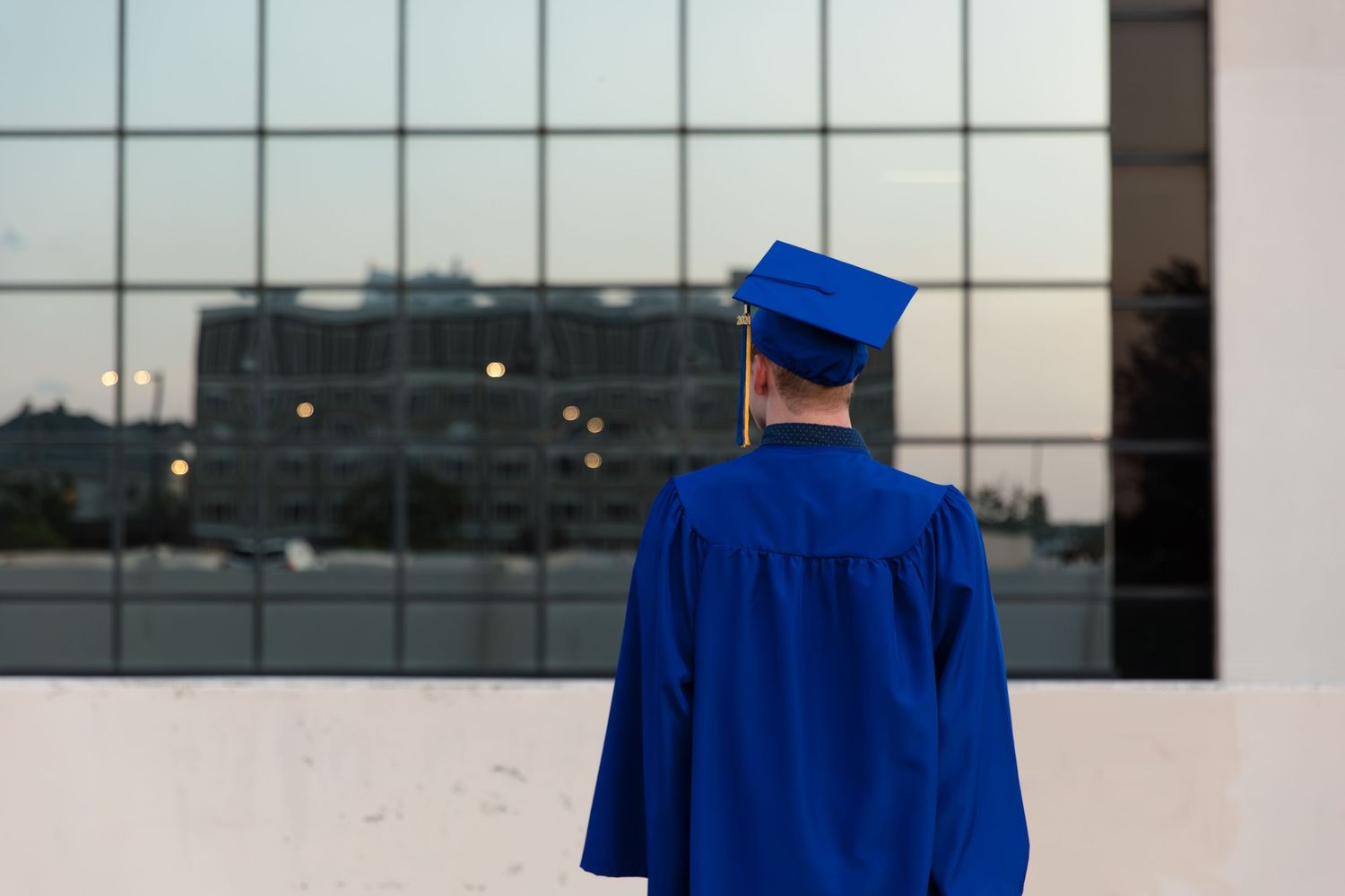 Graduate in blue cap and gown stands facing a large glass building, symbolizing future opportunities.