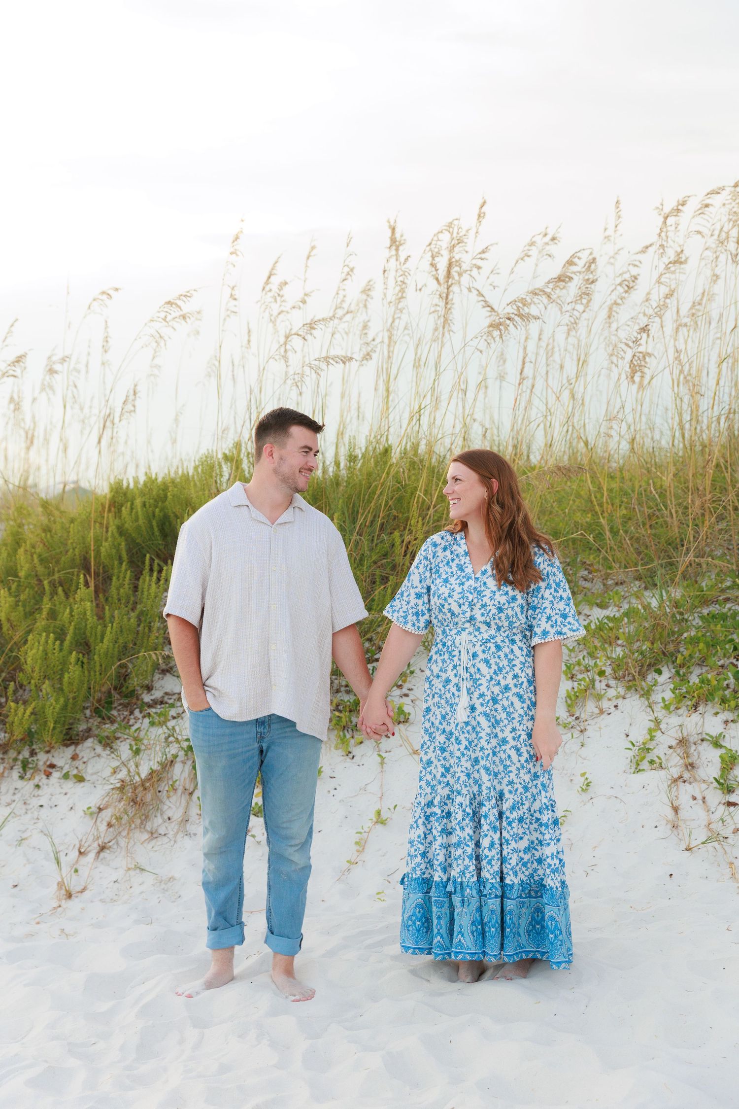 Couple holding hands on white sand beach with sea oats, woman in blue floral maxi dress, man in white shirt and jeans.