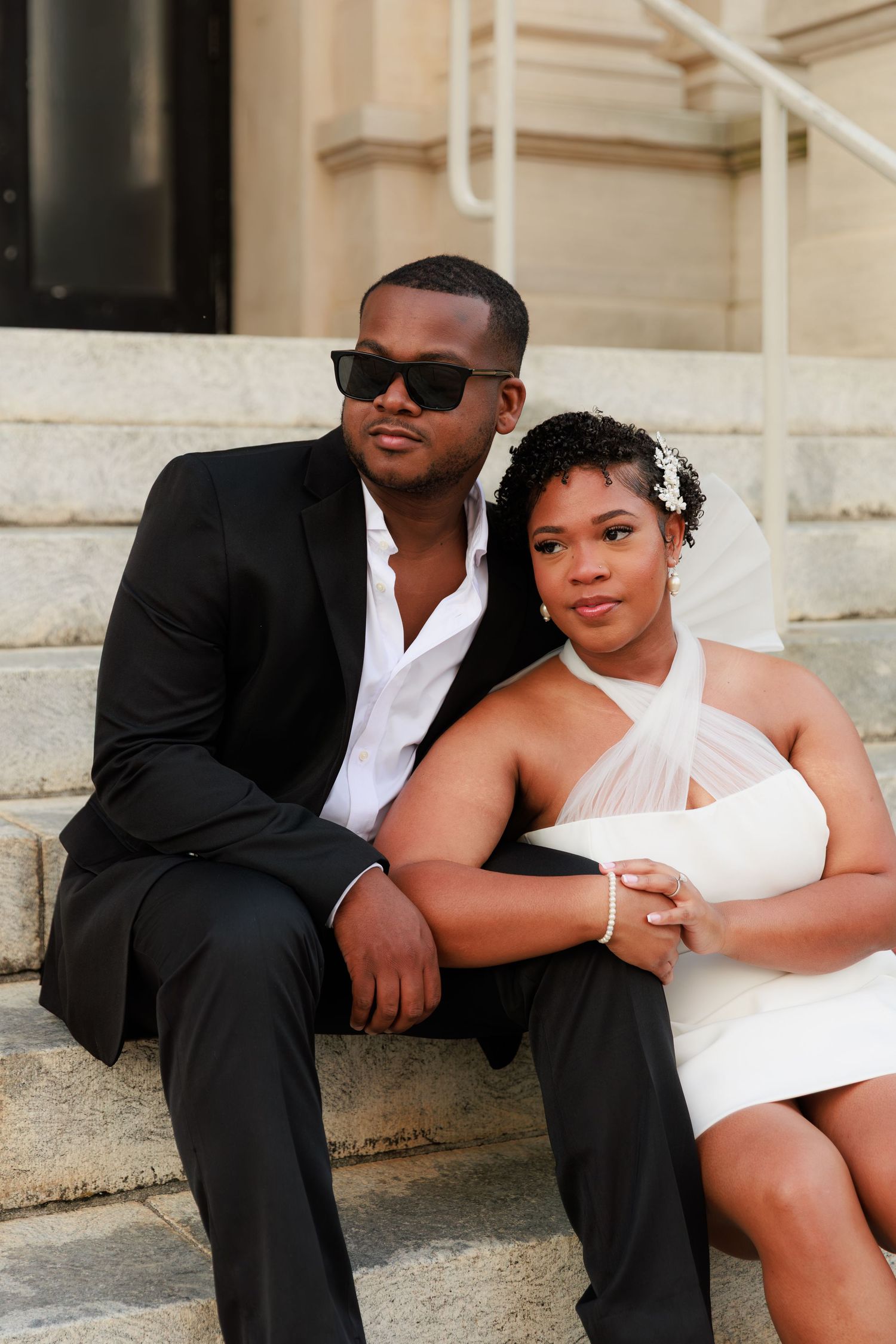 A couple in wedding attire sits on stone steps, groom in black suit and sunglasses, bride in white halter dress with veil.