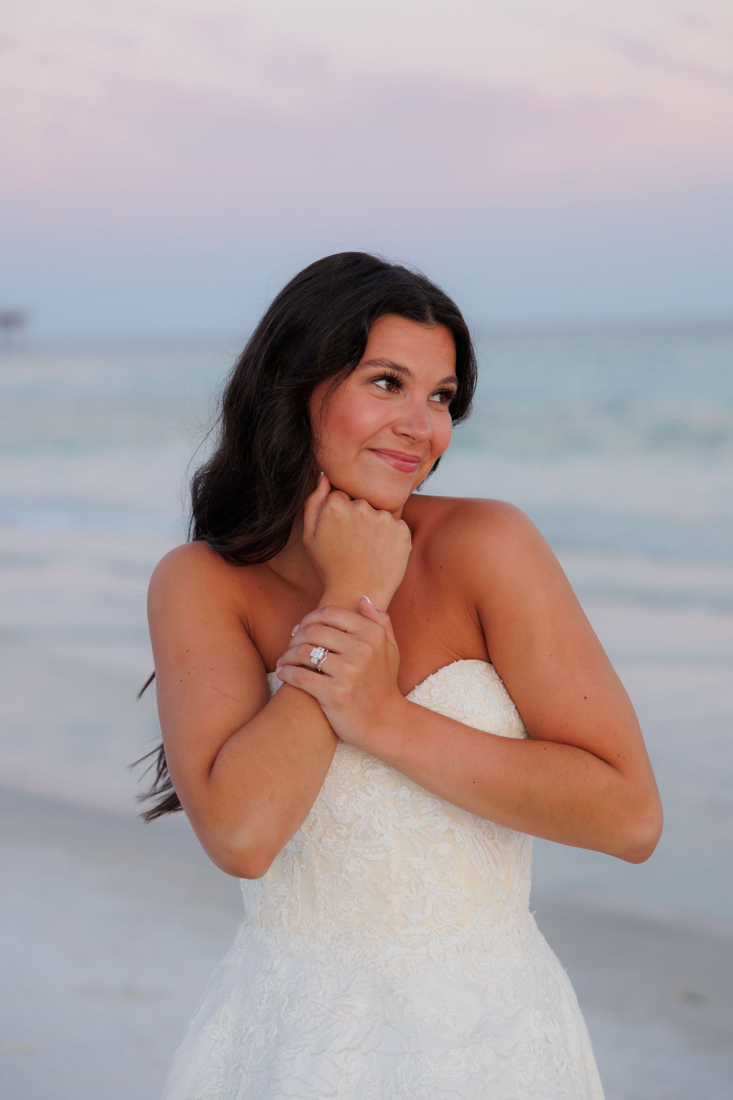 Smiling brunette bride in strapless lace wedding dress poses on beach at sunset.
