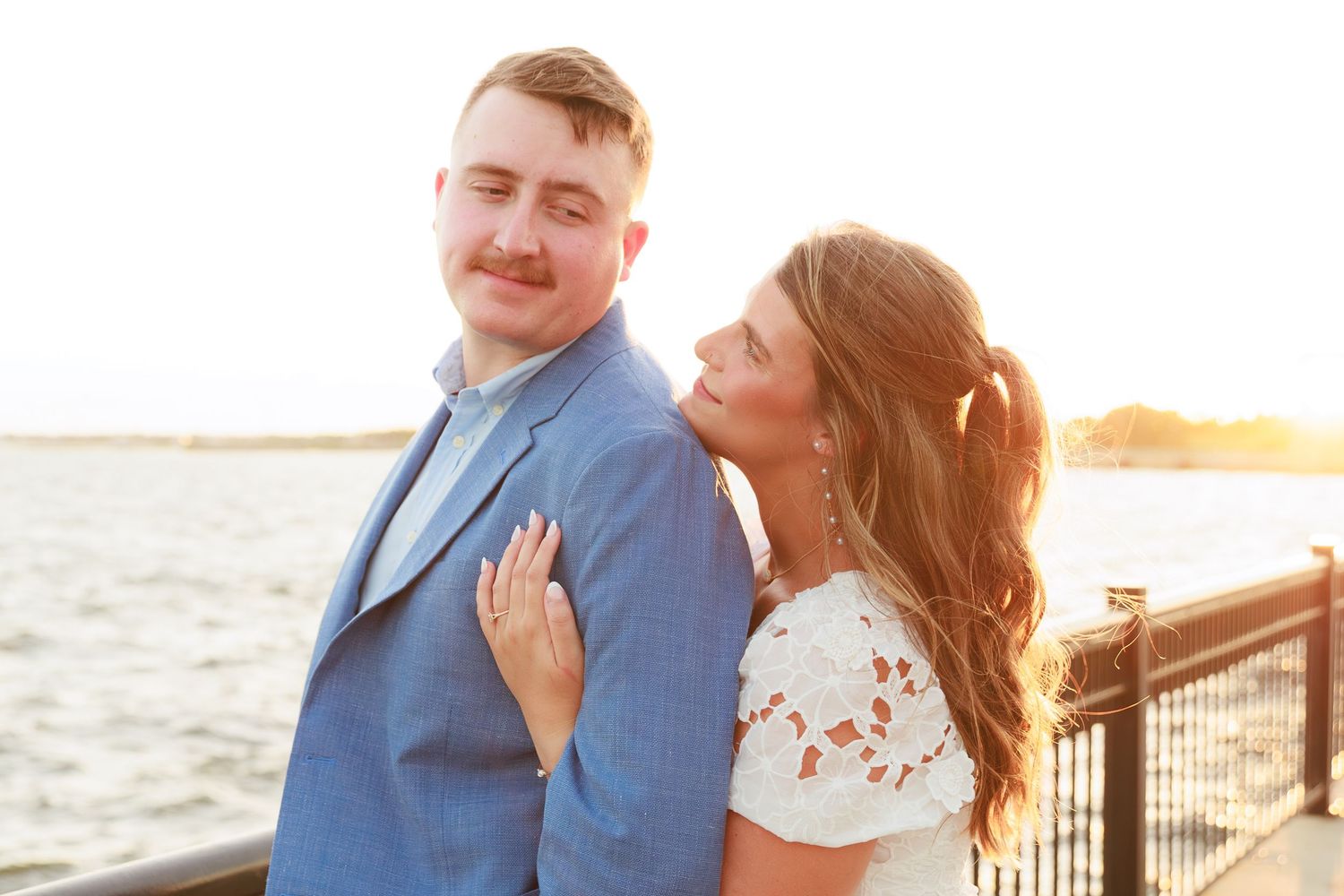 Couple posing at sunset by the waterfront, man in blue suit, woman in white dress hugging him from behind.