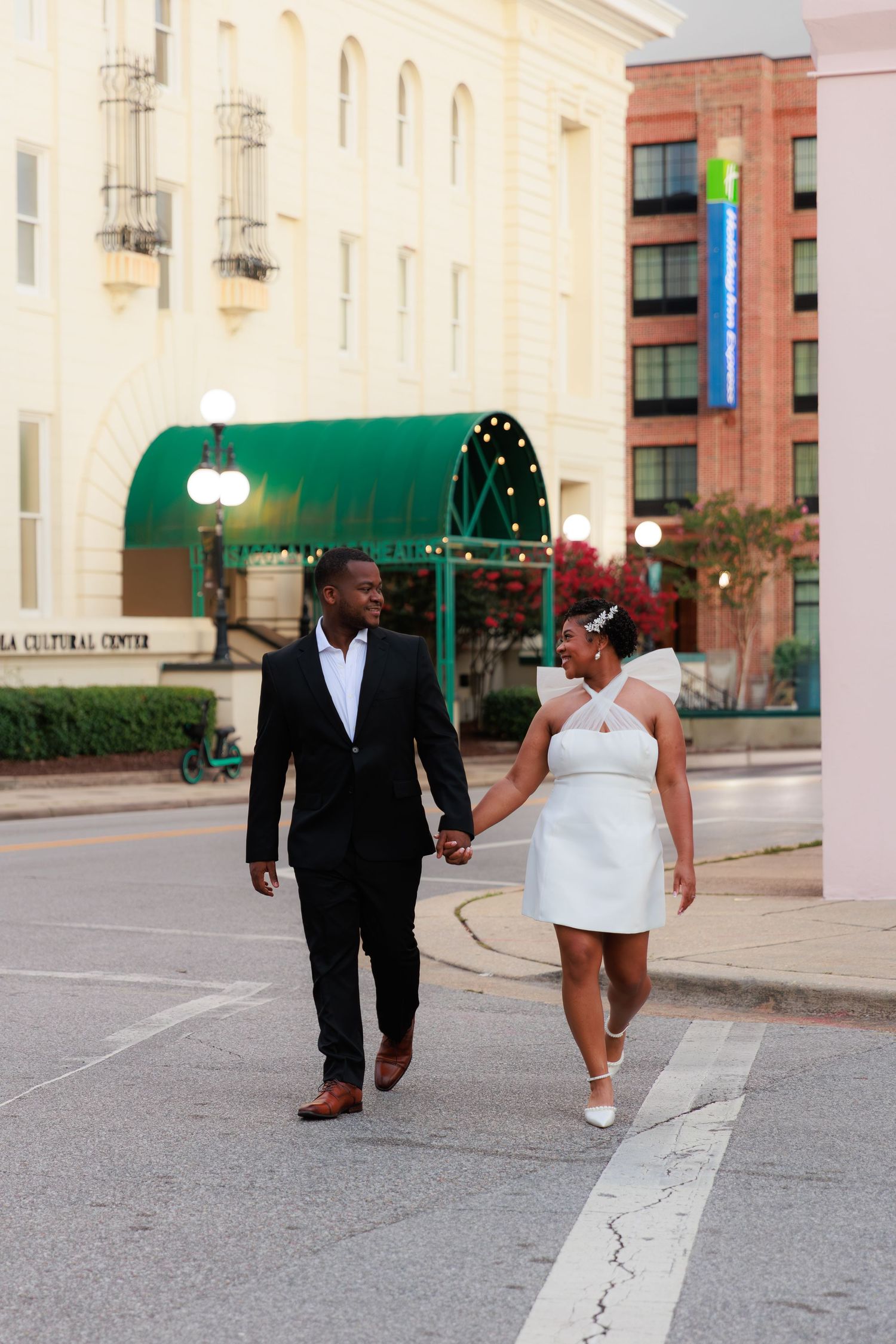 A newlywed couple holds hands while walking across a city street, dressed in a black suit and white mini wedding dress.