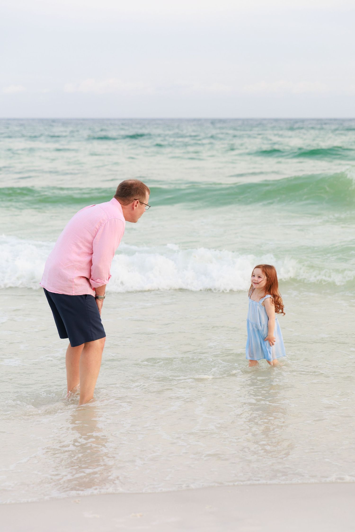 Two people play together in shallow ocean waves on a beach with turquoise water and overcast sky.