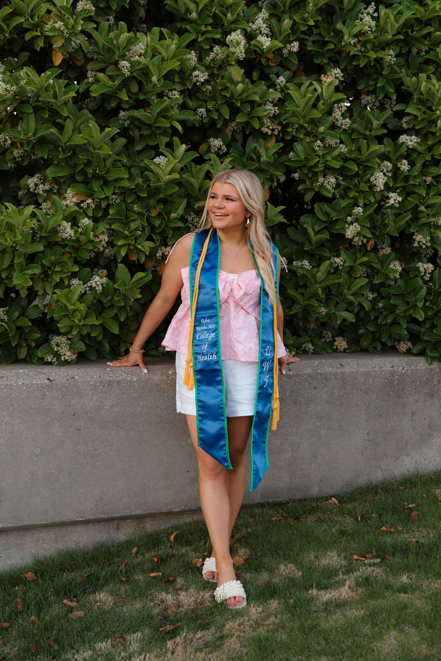 Graduate in pink dress and blue graduation stole poses in front of flowering bush on sunny day.